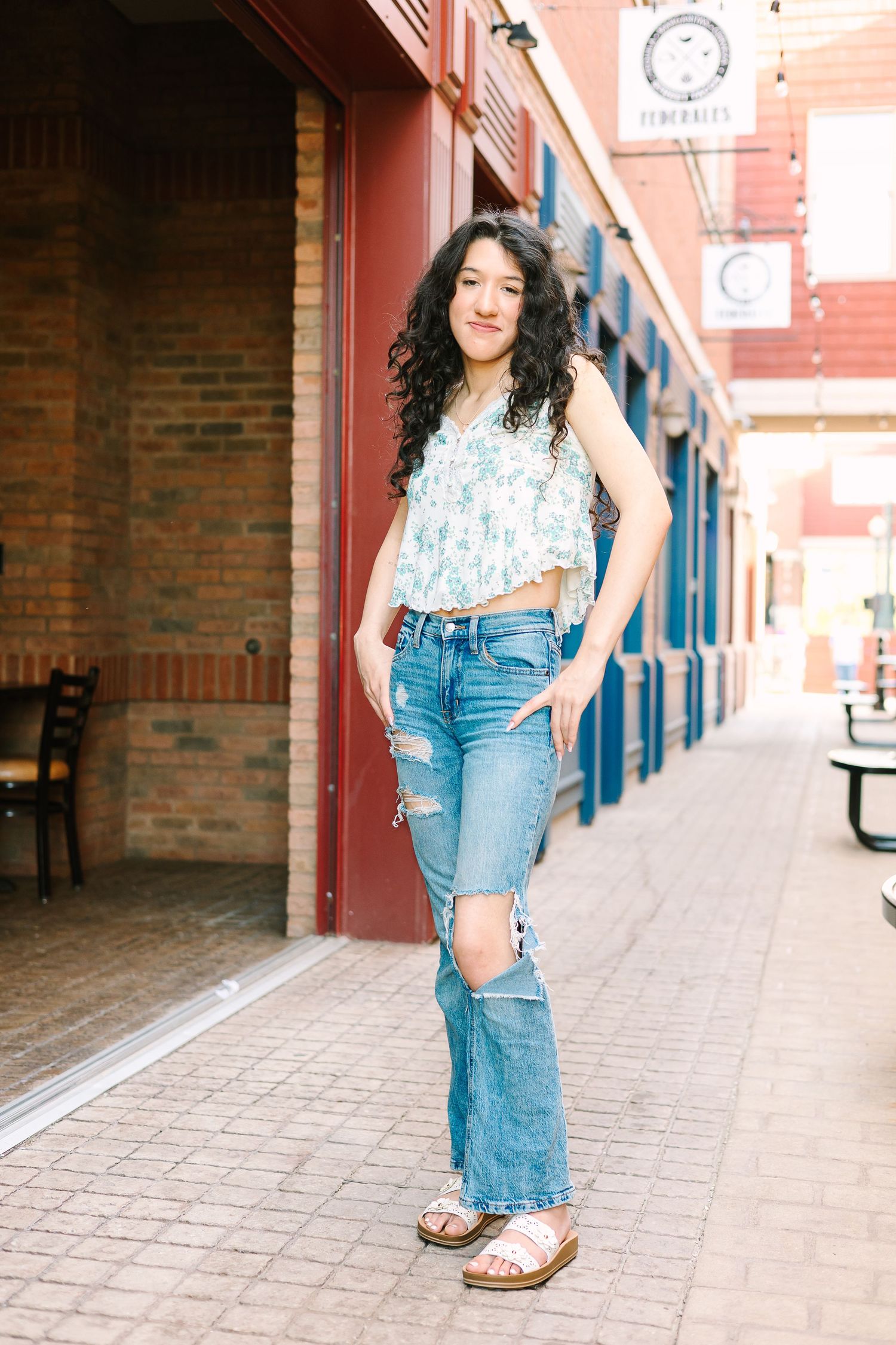 A casual summer outfit featuring ripped jeans, a floral top and sandals against a brick storefront background.