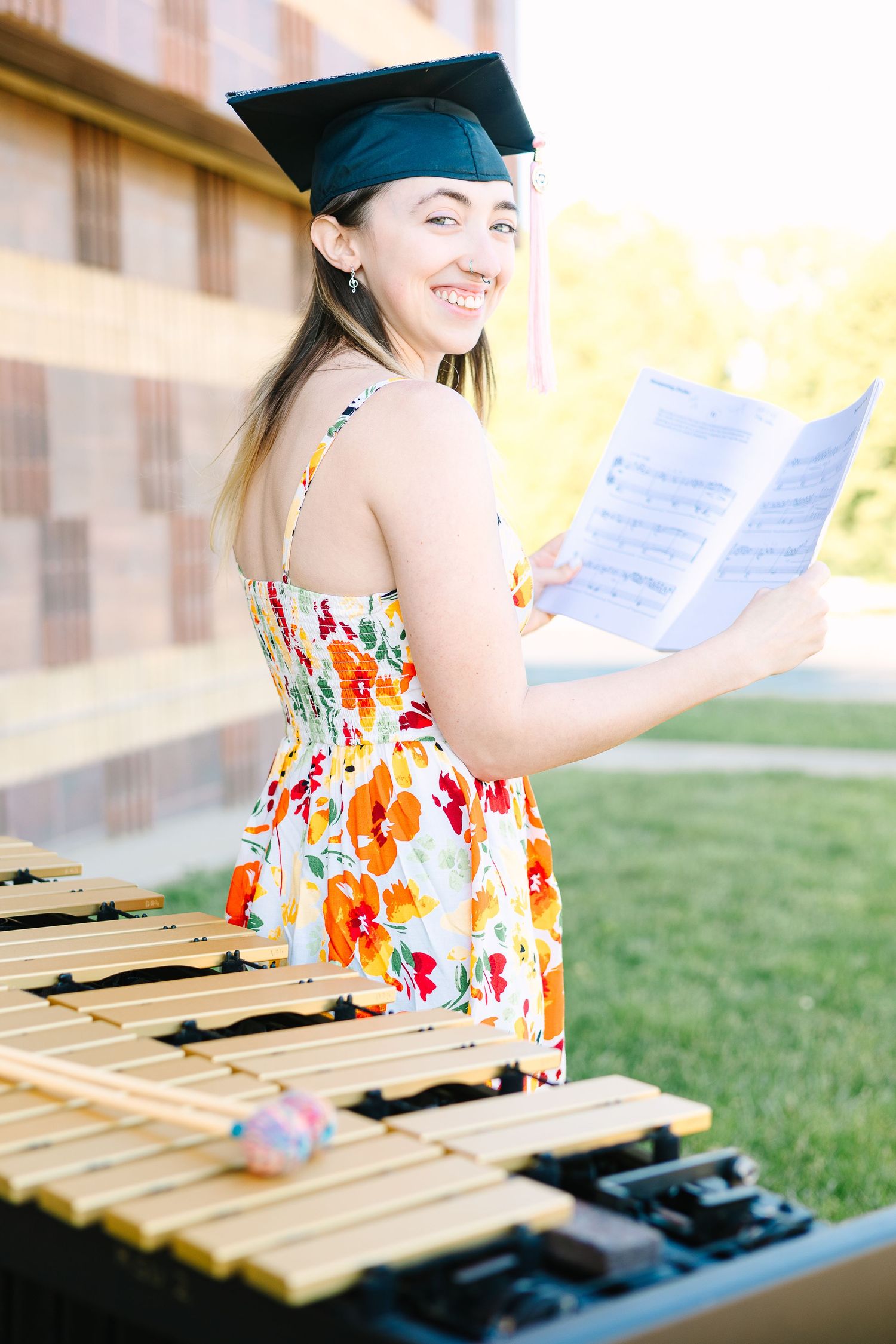 A smiling graduate in a colorful floral dress plays the xylophone on a sunny day outdoors.
