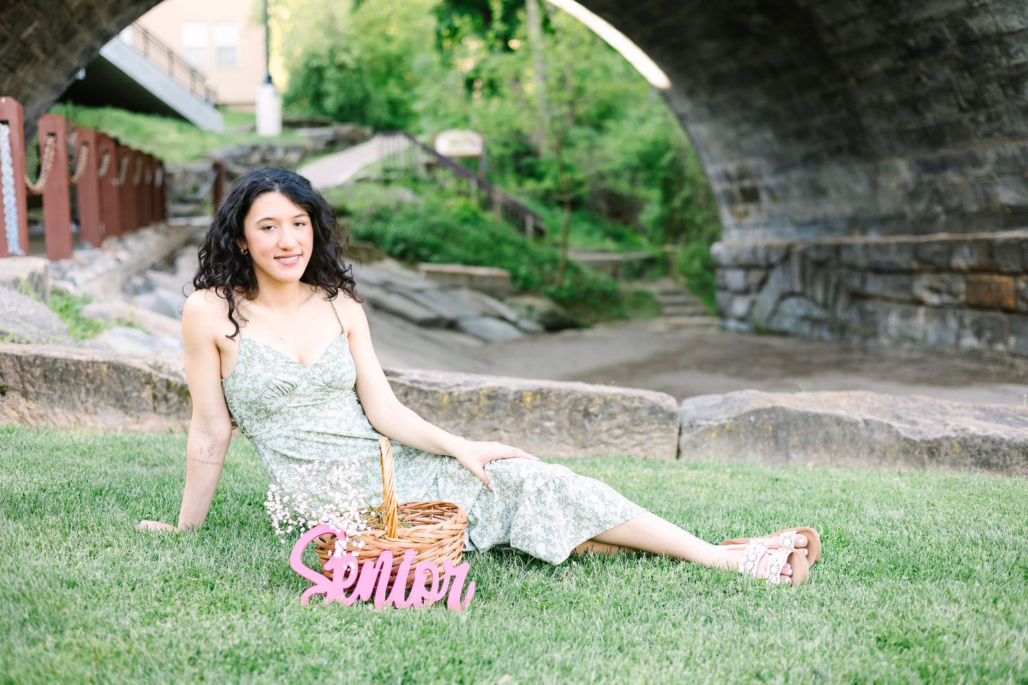Someone in a floral sundress lounges on green grass with a picnic basket on a sunny day near stone steps.