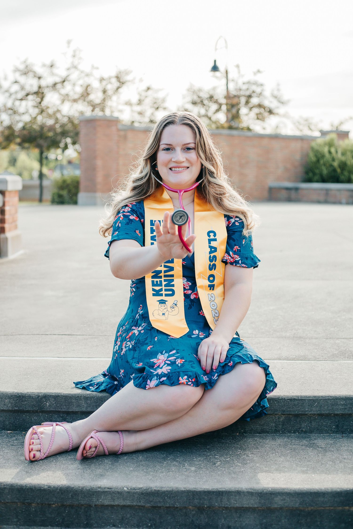 Graduate in blue floral dress sits on steps wearing academic stoles.