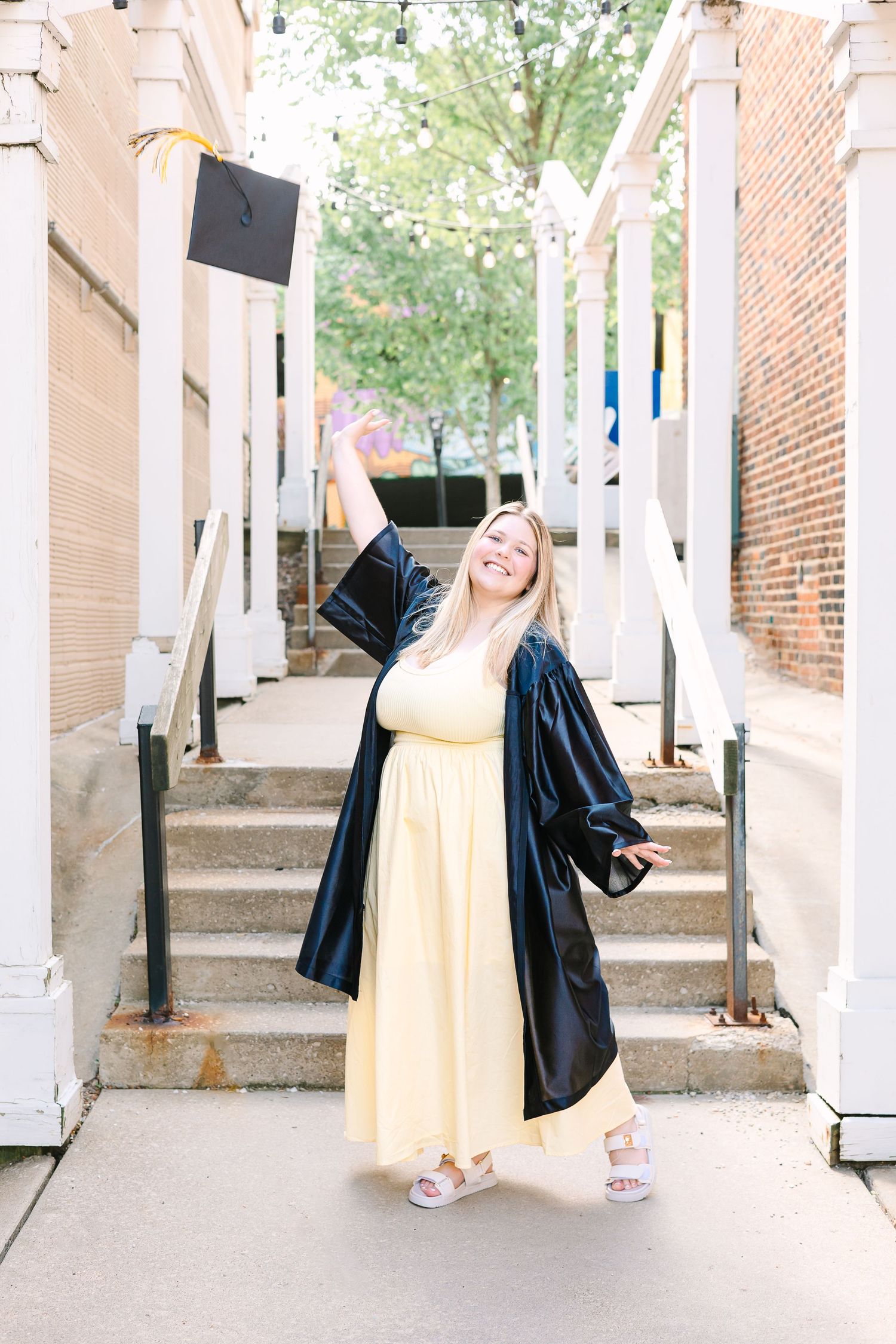 Graduate in yellow dress and black graduation gown poses joyfully on outdoor campus steps.