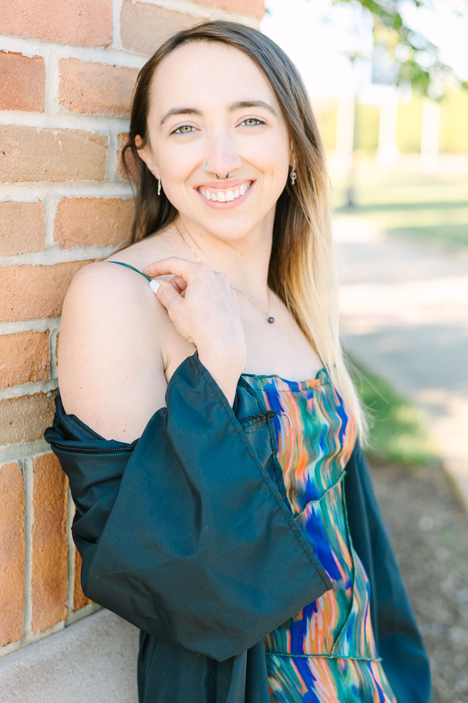 Smiling person wearing colorful patterned dress and dark cardigan poses against brick wall in outdoor setting.