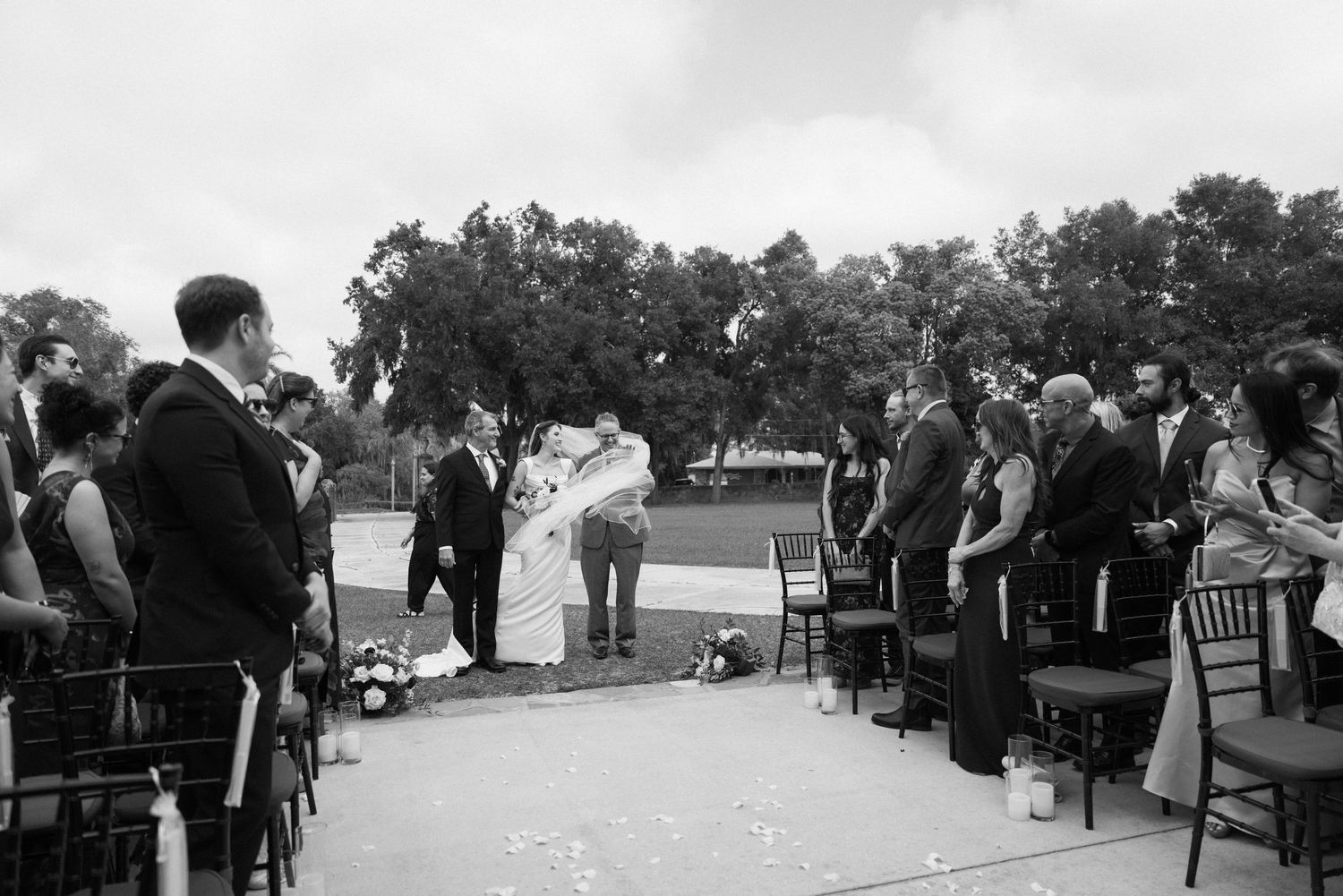 Black and white outdoor wedding ceremony with couple at altar and seated guests on lawn.