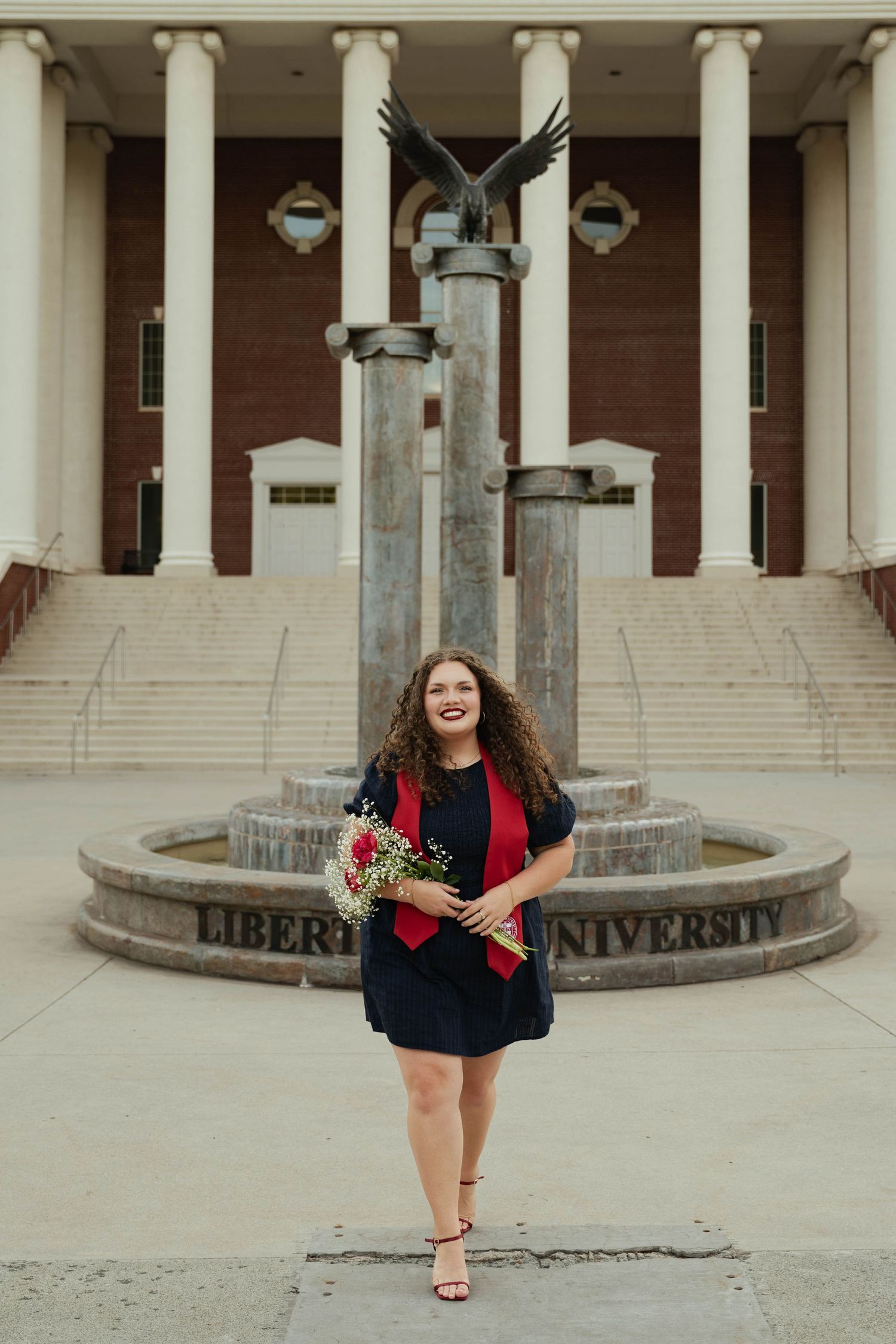 Smiling graduate in navy dress holds bouquet in front of Liberty University fountain and eagle statue.