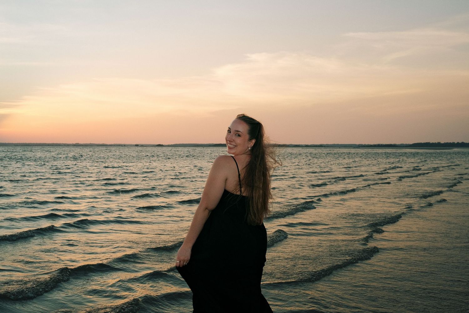 Woman in black dress smiling at sunset beach, hair blowing in wind with ocean waves behind her.
