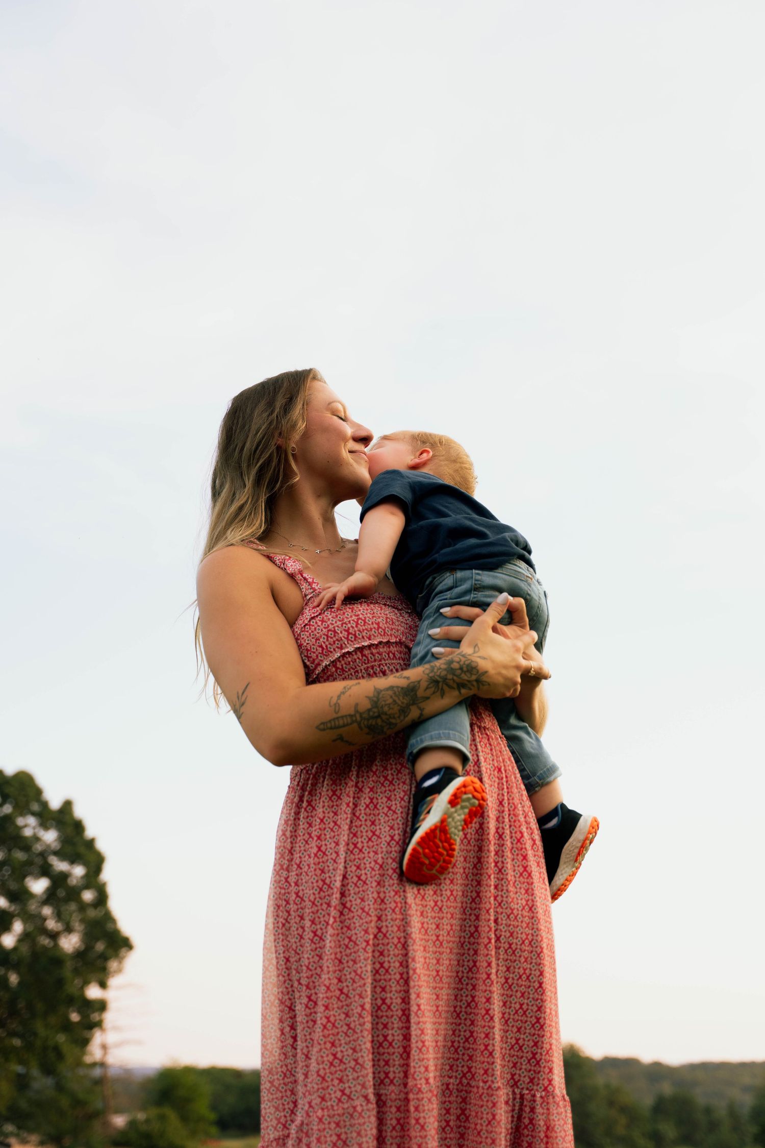 Woman in a pink floral dress holds and lifts a small child playfully outdoors.