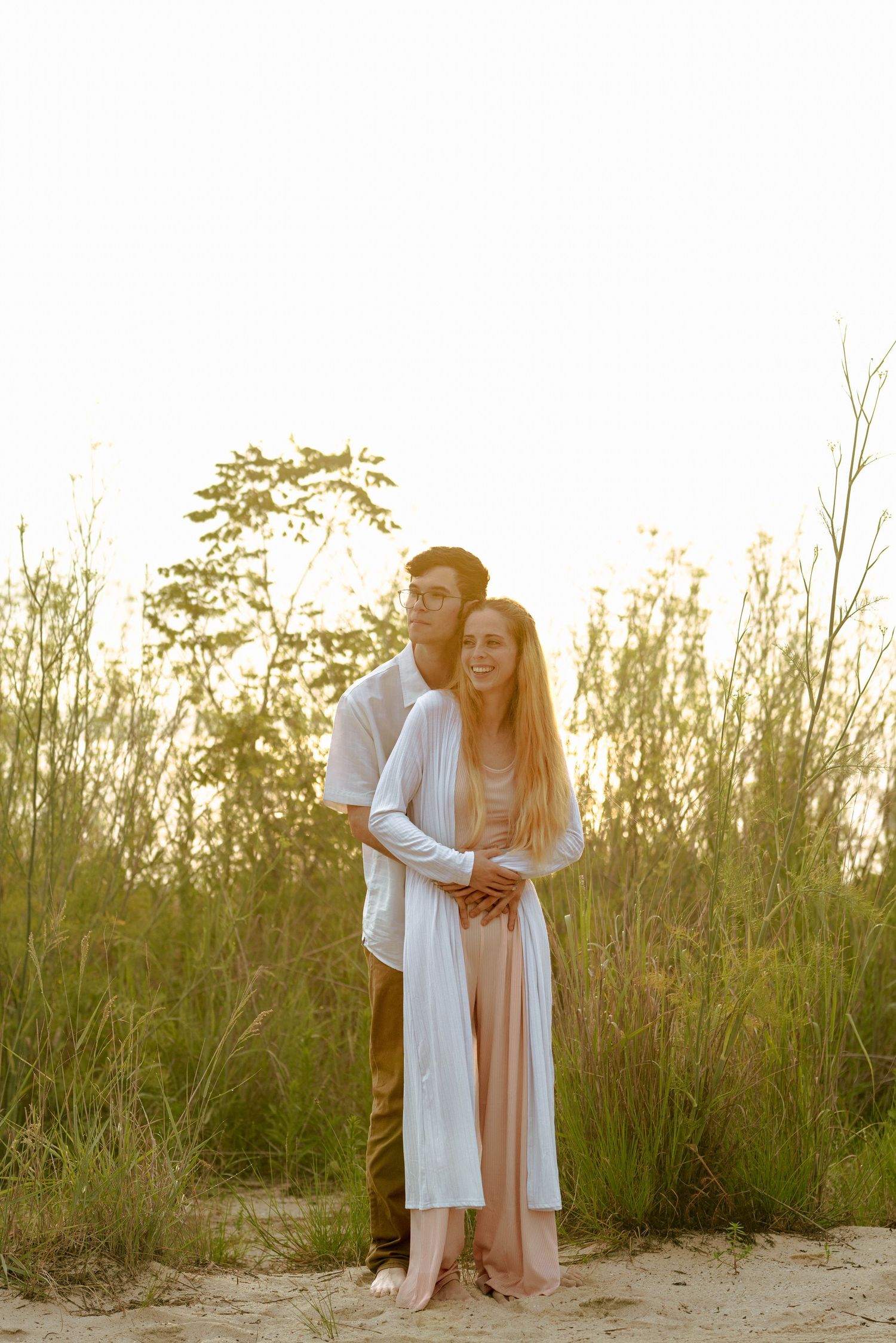 A couple in light colored clothing shares a tender moment in tall grass at sunset.