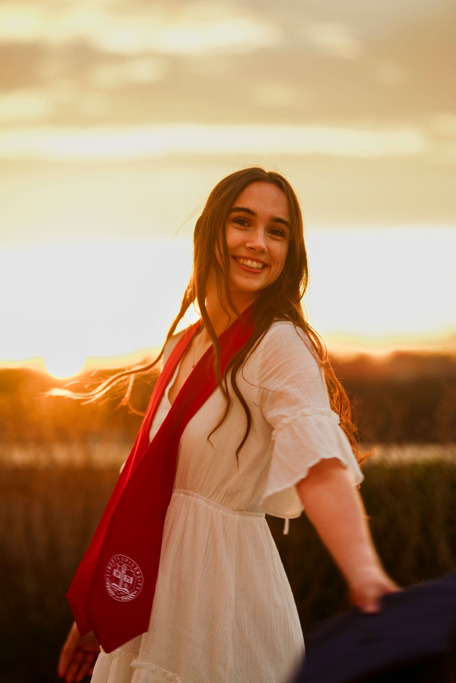 A person in a white dress and red scarf smiles during golden hour sunset outdoors.