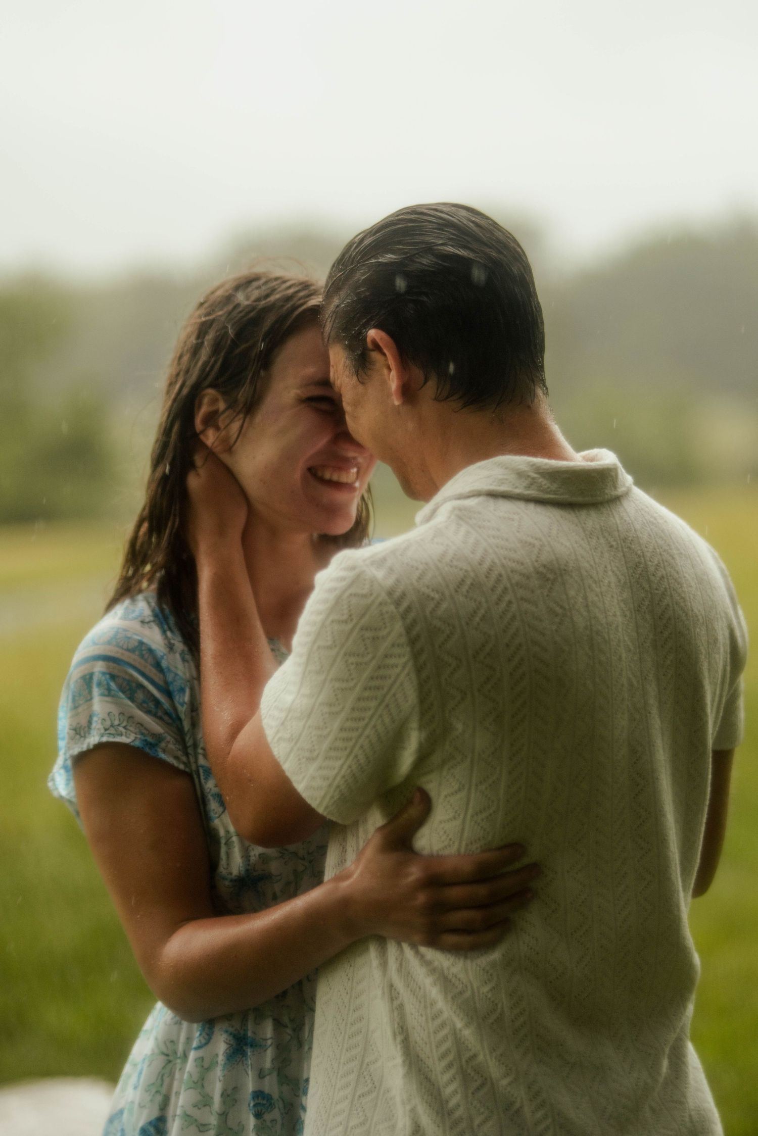 Couple embracing and smiling in the rain, soaking wet, sharing a romantic moment outdoors in a green field.