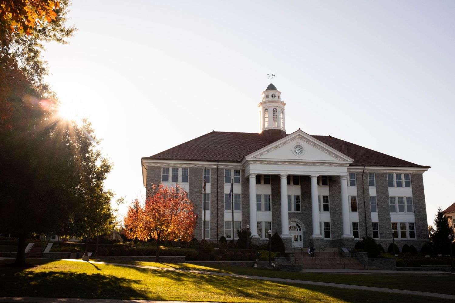 A stately white building with a clock tower stands amidst the vibrant fall foliage on a grassy lawn.