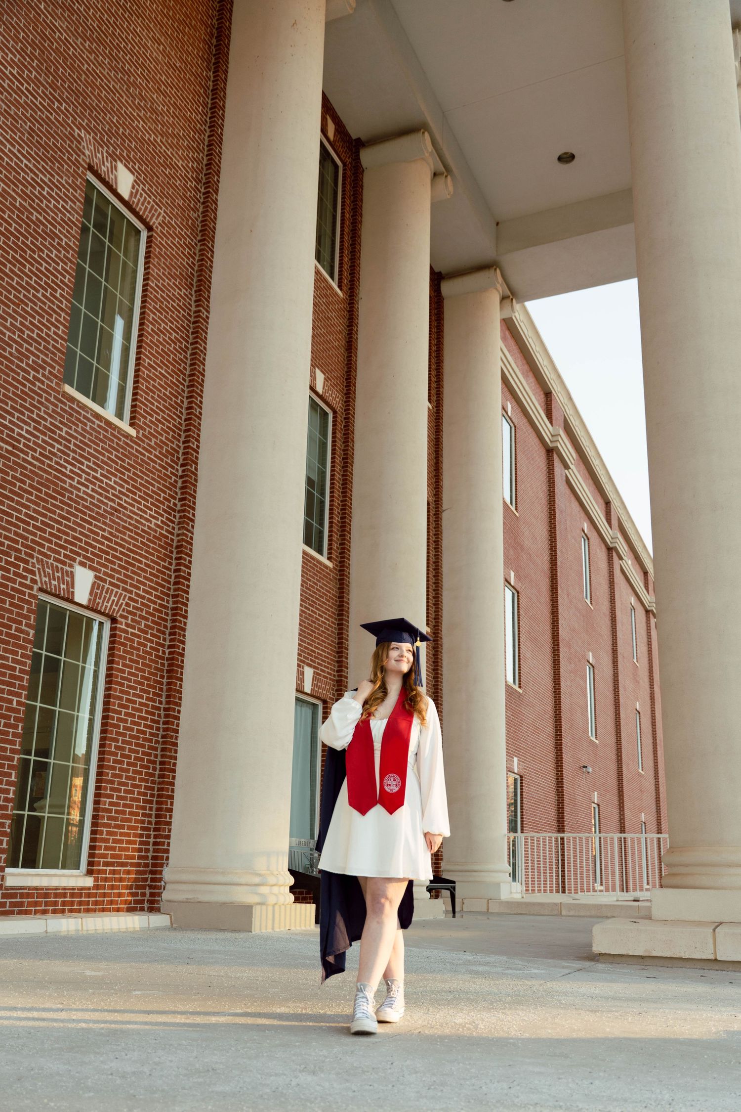 A young woman in a graduation gown and cap stands in front of a brick building, ready to celebrate her academic achievement.