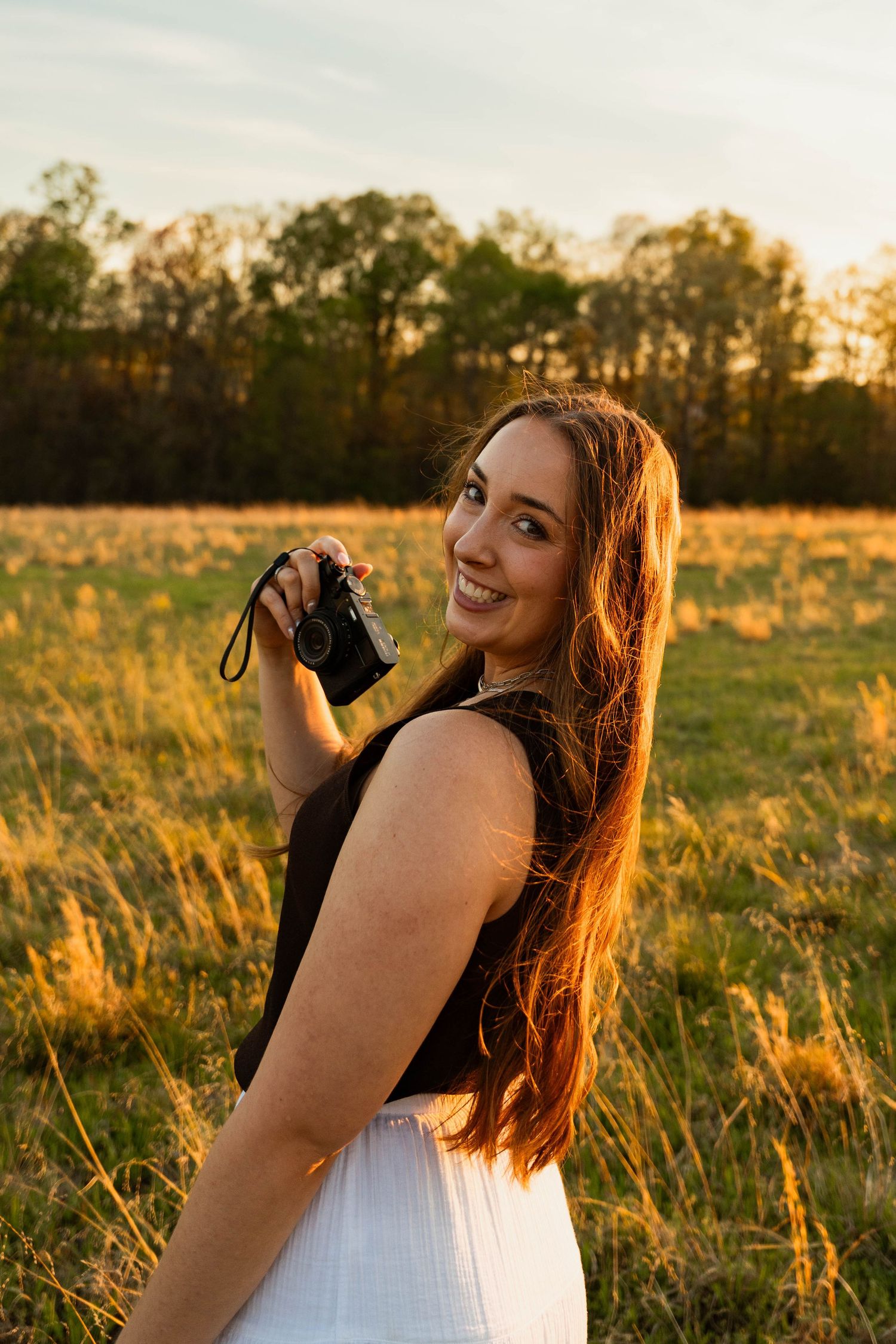 Smiling woman holding a camera in a golden sunset field, wearing a black top and white skirt with long brown hair.