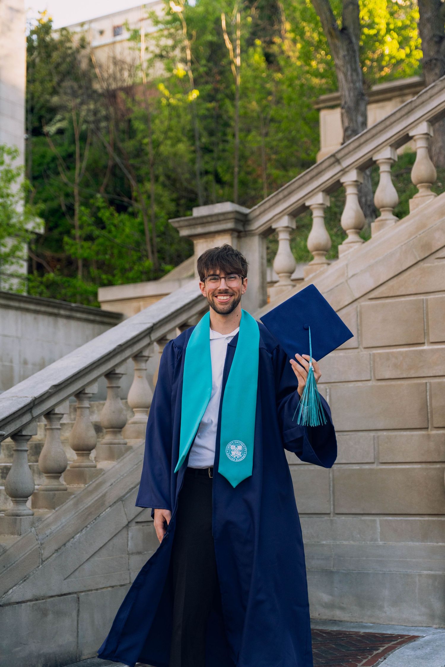 A young man in a blue graduation gown stands on stone steps, holding a book and smiling.