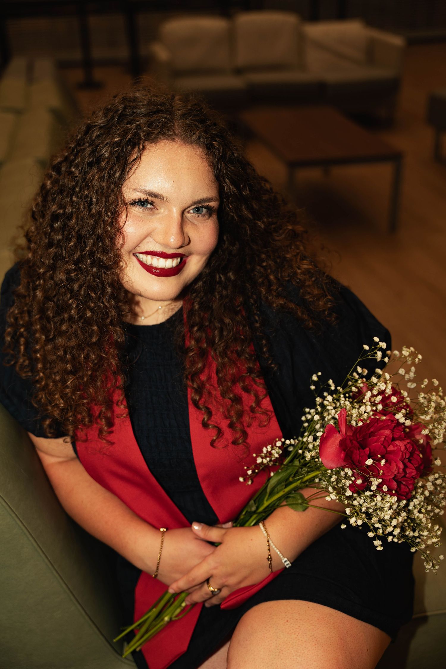 Smiling young woman with curly hair holding red roses and baby's breath bouquet, wearing red and black outfit.