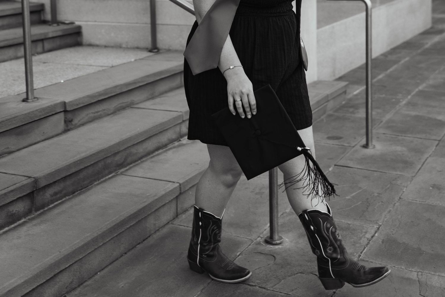Woman wearing black dress and fringed cowboy boots walking on stone steps in black and white photo.