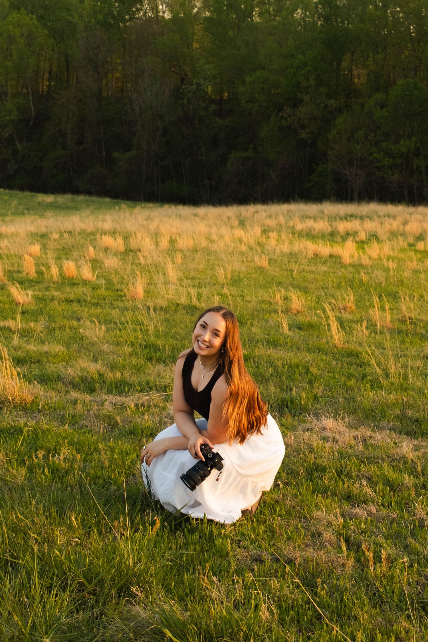 Young woman with long red hair crouches in a sunlit meadow, smiling while holding a camera, with green trees in background.