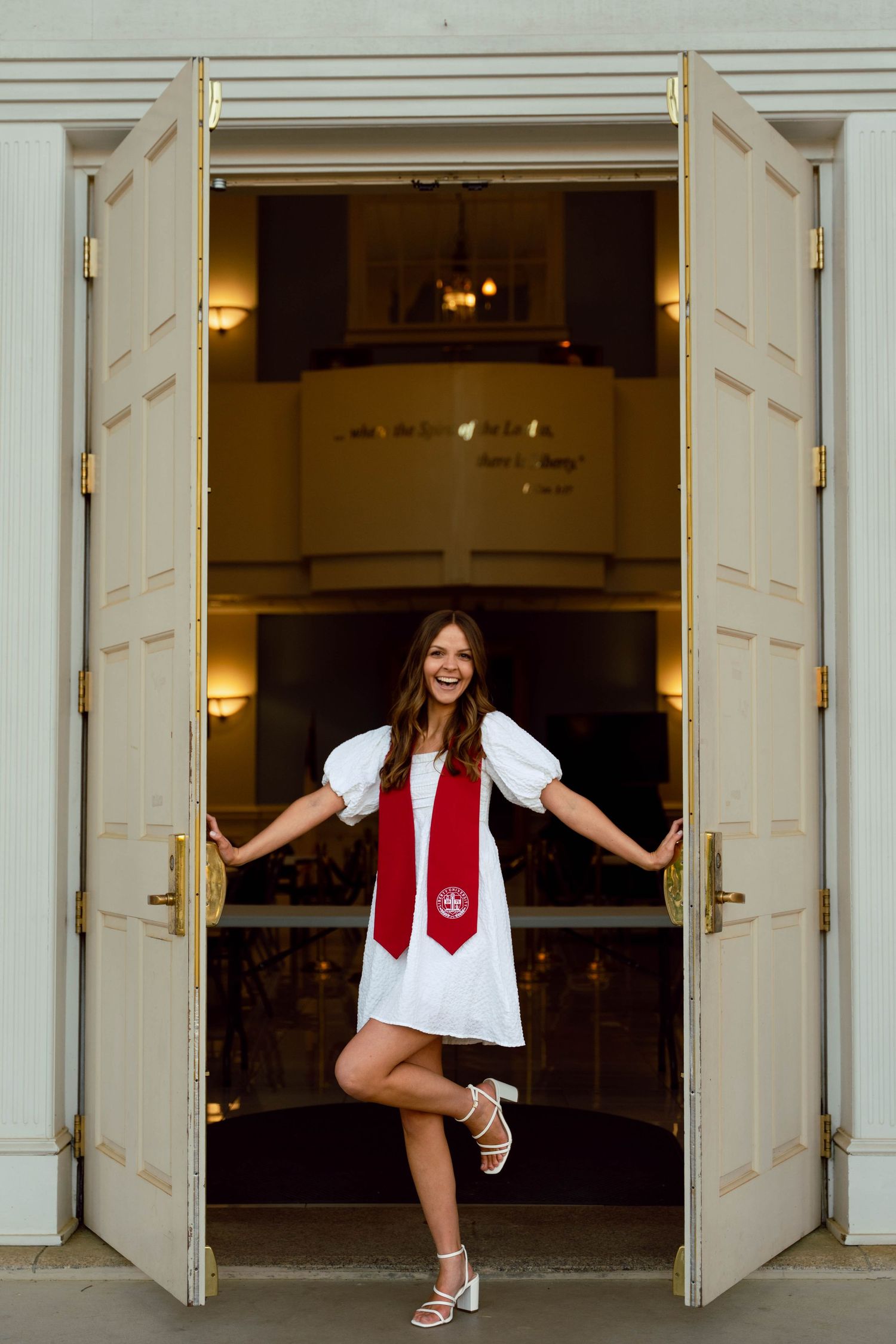 Graduate in white dress and red stole poses playfully between white columns.