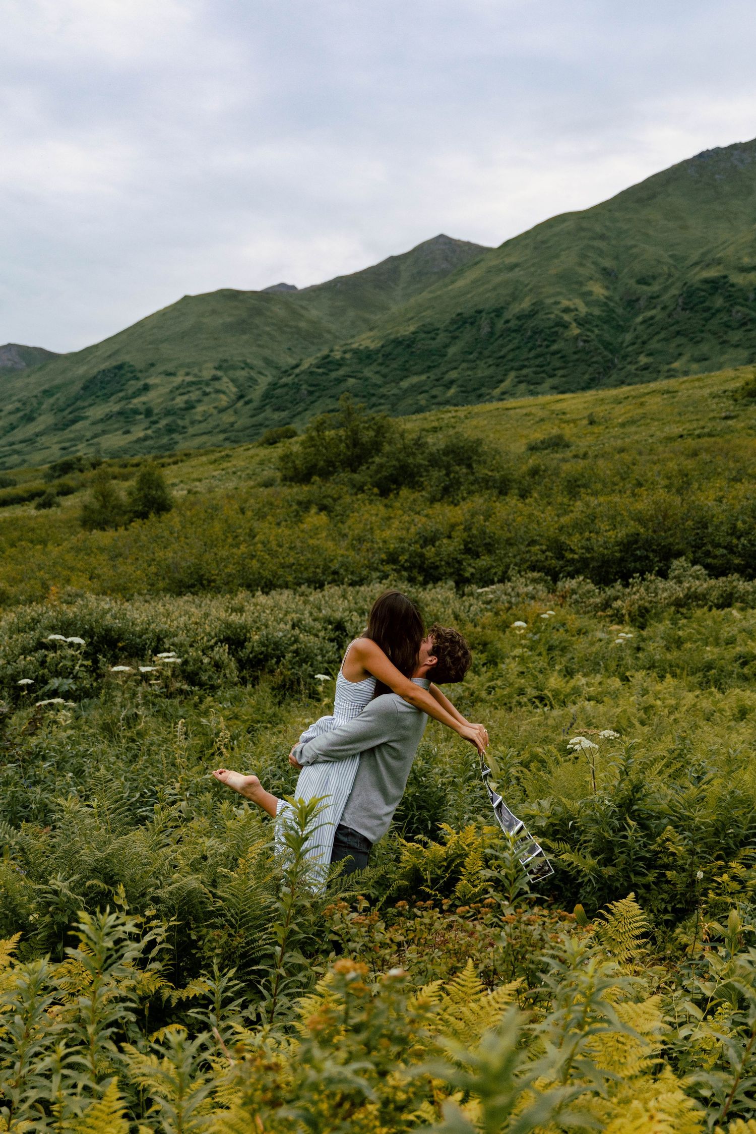 Couple embracing in lush green mountain meadow with rolling hills and overcast sky in background.