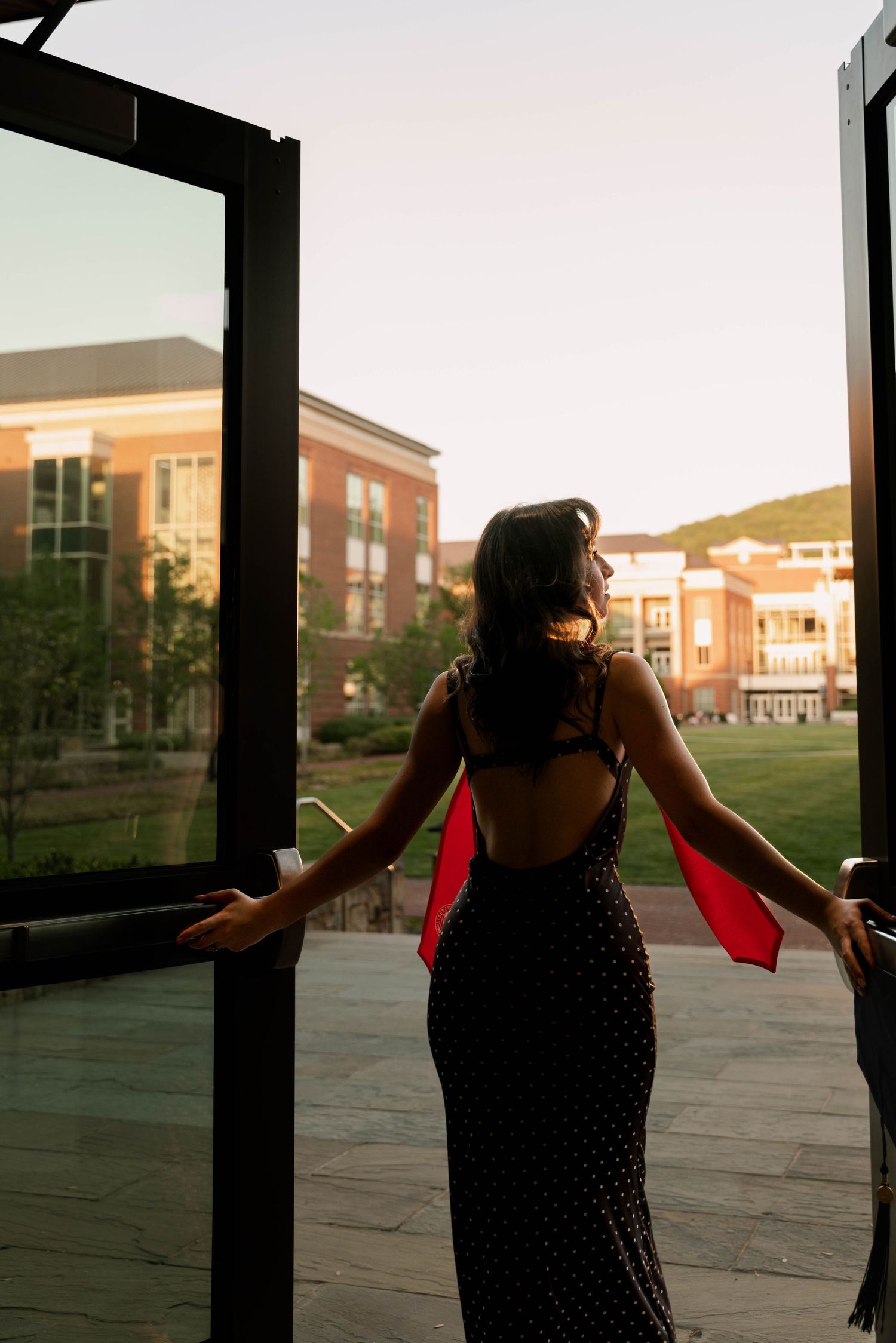 Woman in backless black polka dot gown opens glass doors to a brick university campus at sunset.