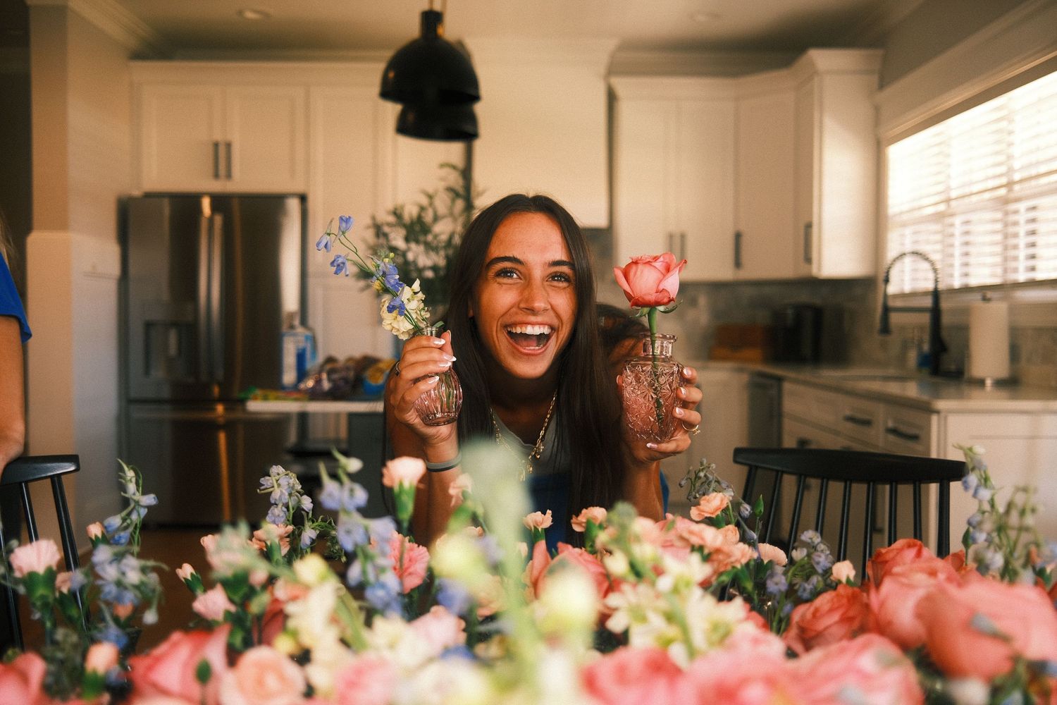 A smiling woman surrounded by flowers and holding a glass of wine in a kitchen.