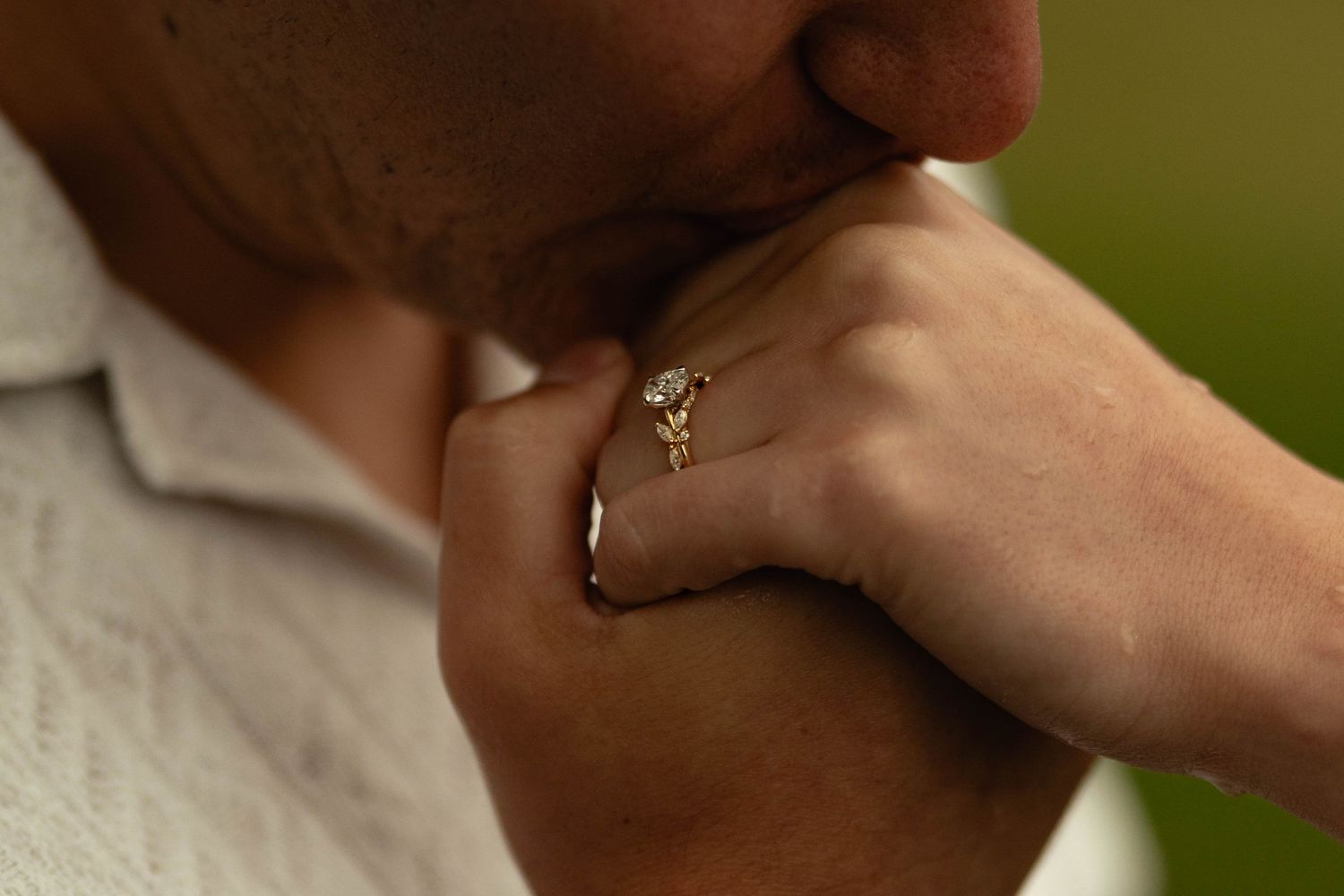 Close-up of a person kissing a hand adorned with a gold diamond engagement ring against a blurred green background.