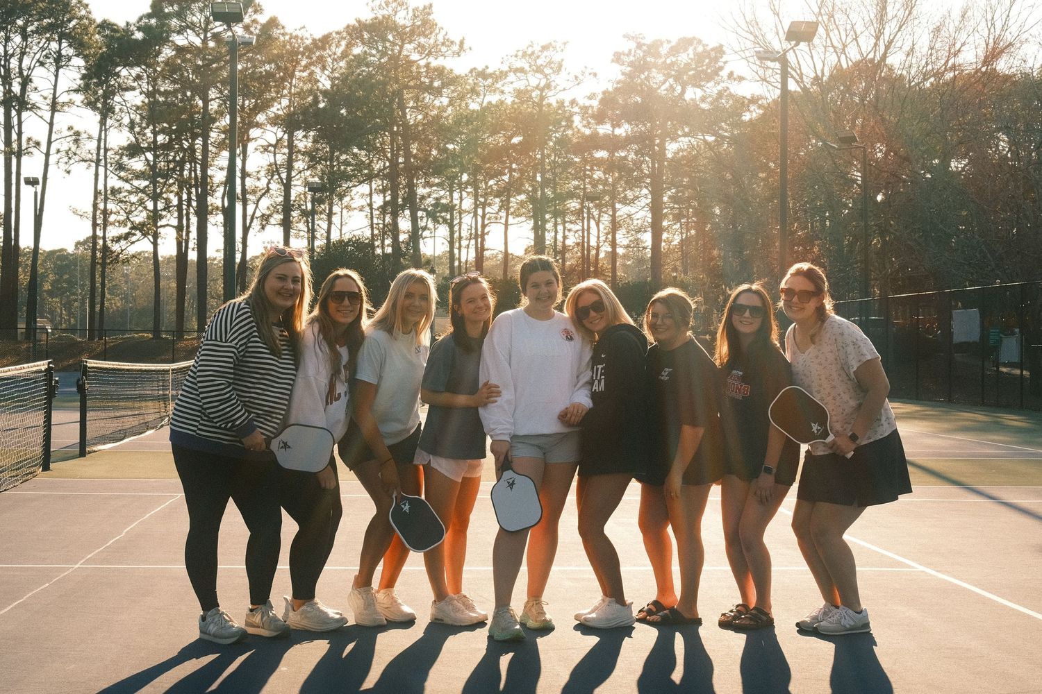 A group of young women posing together outdoors in a park setting with trees and sunlight.