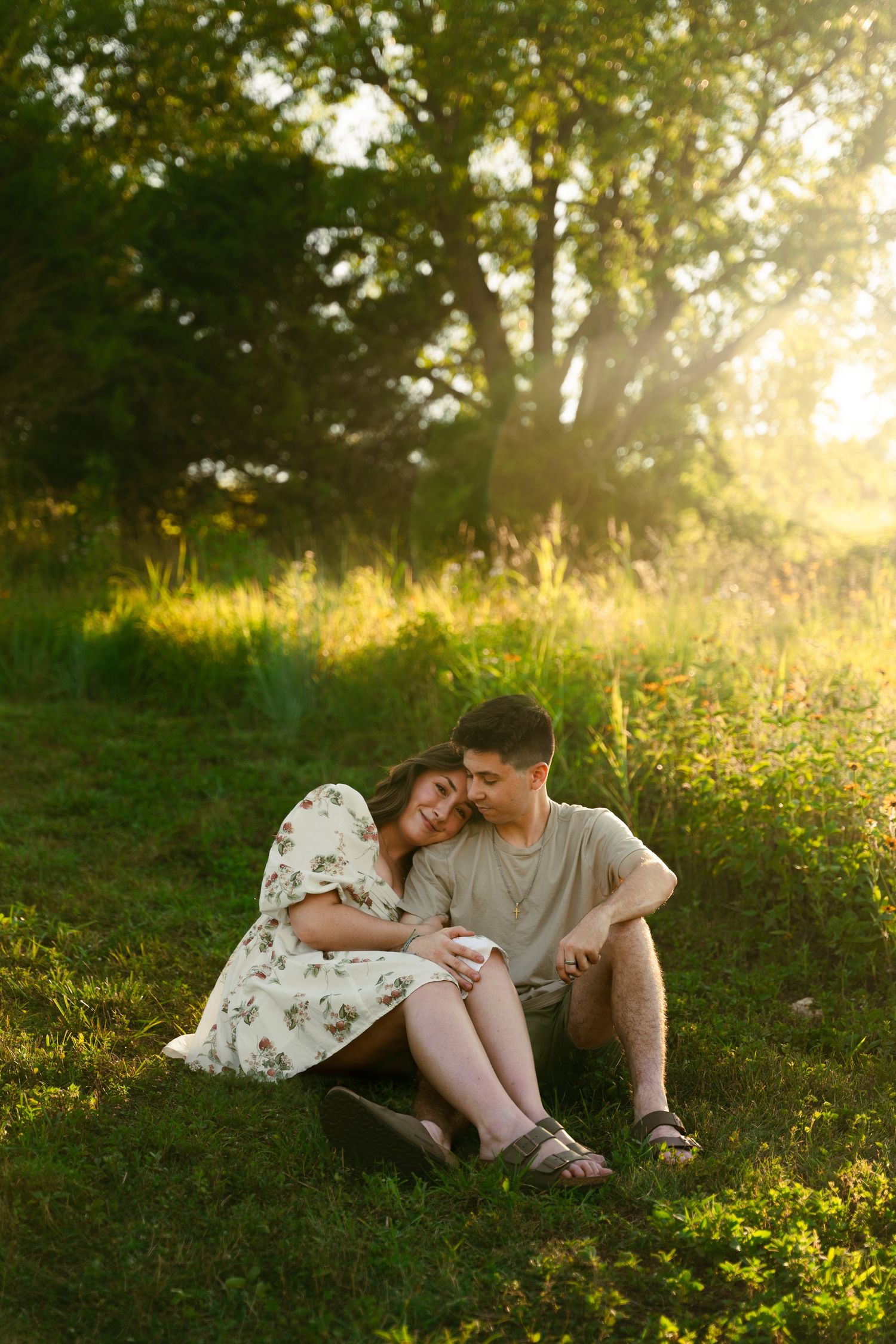A young couple sitting on the grass in a lush, sunlit forest setting.