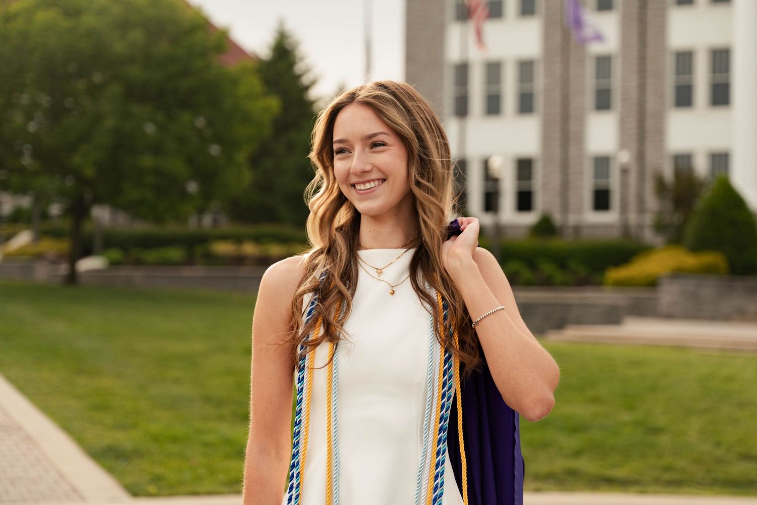 Smiling graduate in white dress holding purple gown with colorful honor cords outdoors near brick building.