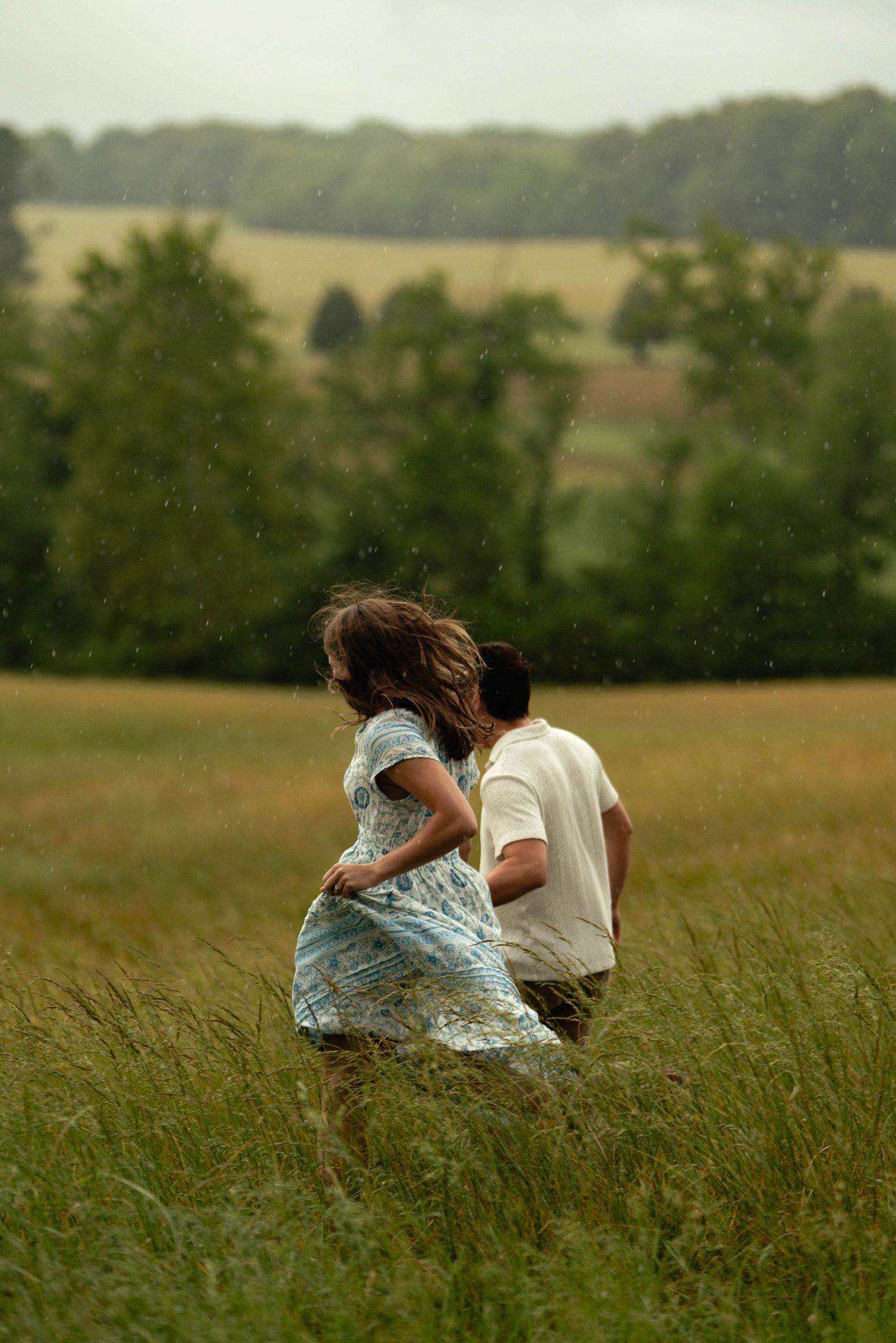 A couple runs through a lush green field in the rain, woman in blue dress with flowing hair, trees in background.