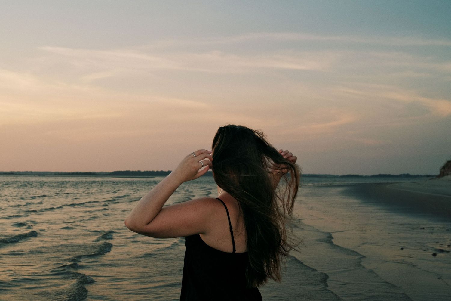Woman with long dark hair standing on a beach at sunset, holding her hair in the breeze.