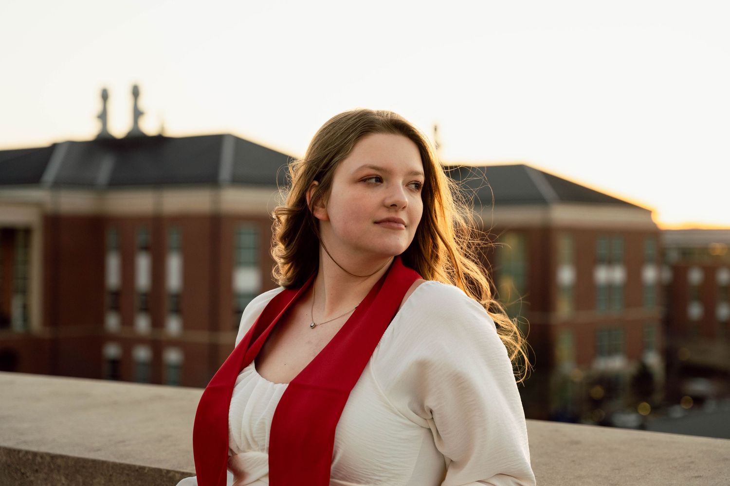 Graduate in white dress and red stole poses during sunset near brick buildings.