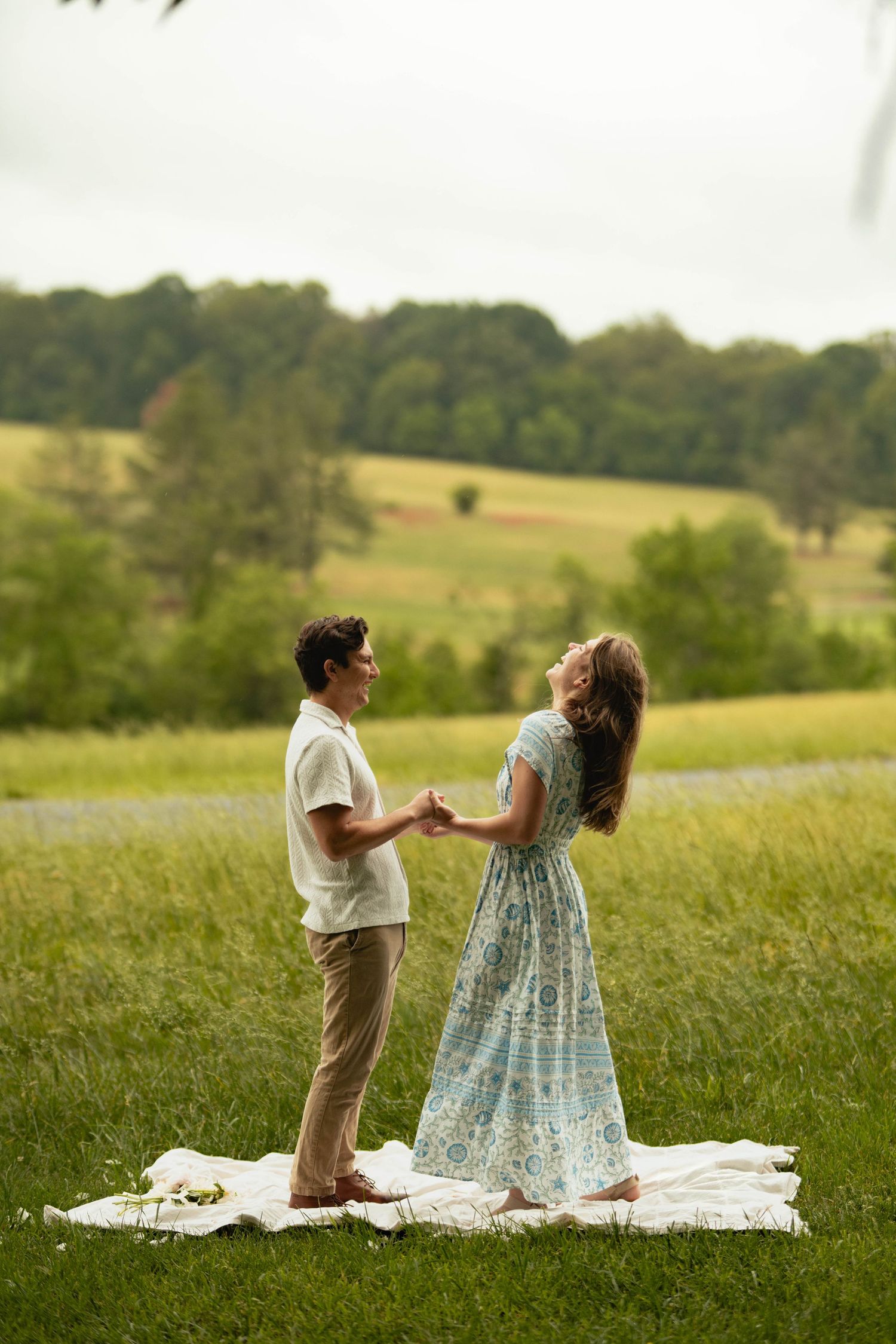 A couple holds hands and dances on a white blanket in a lush green meadow, the woman wearing a flowy blue floral dress.