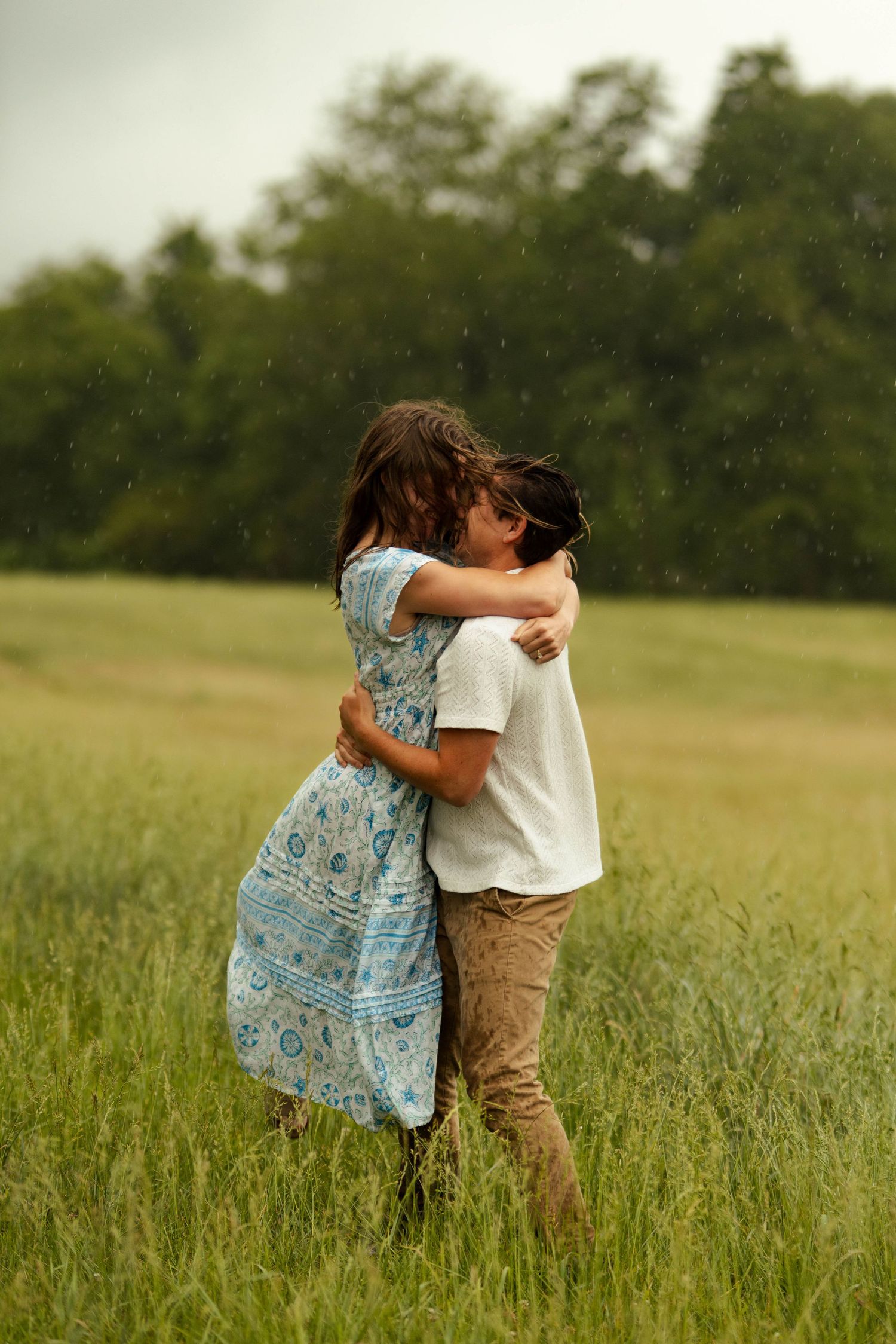 Couple embracing romantically in a green field during rain, woman in blue floral dress and man in white shirt.
