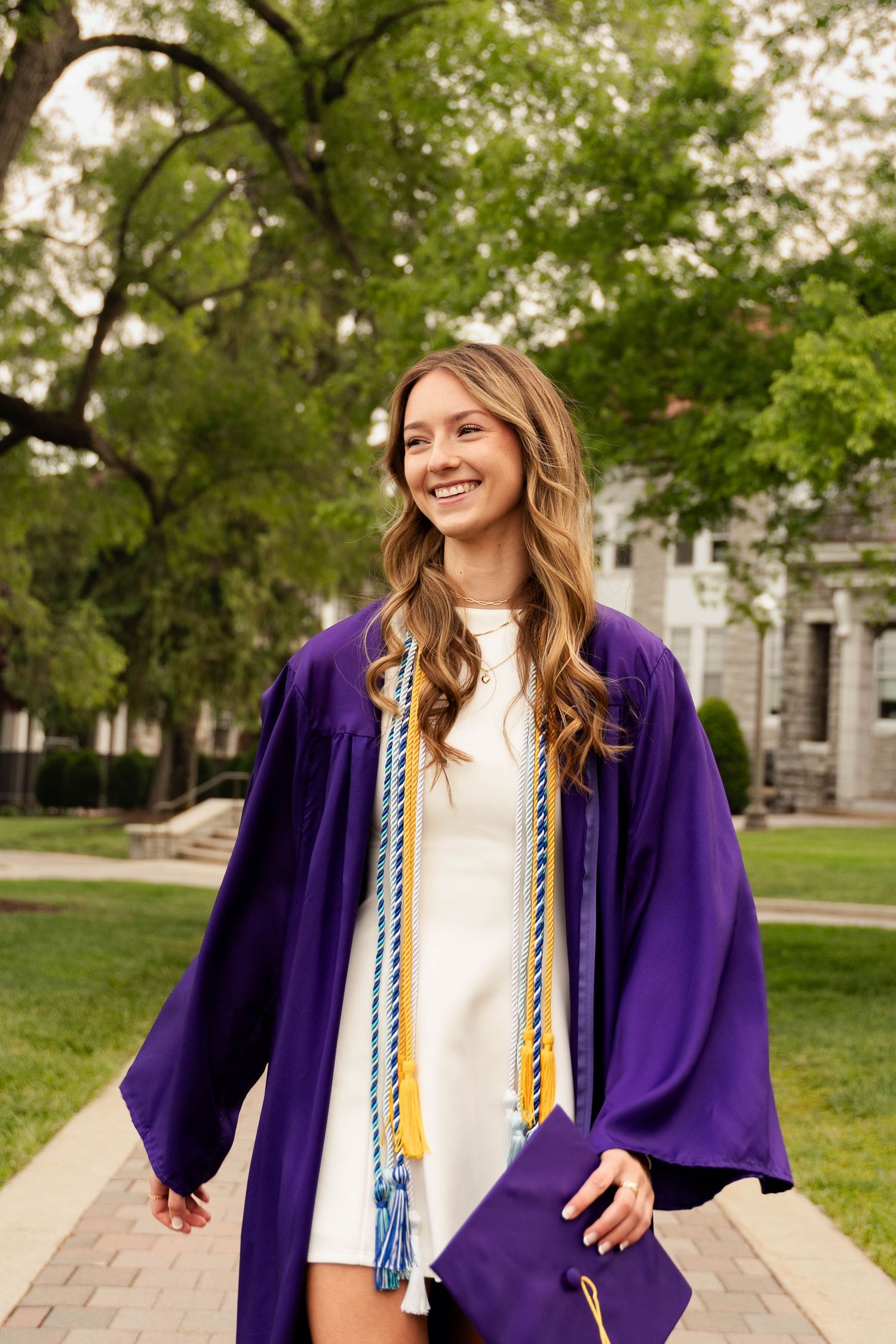 Smiling young woman in purple graduation gown and honor cords holding cap on a tree-lined campus walkway.