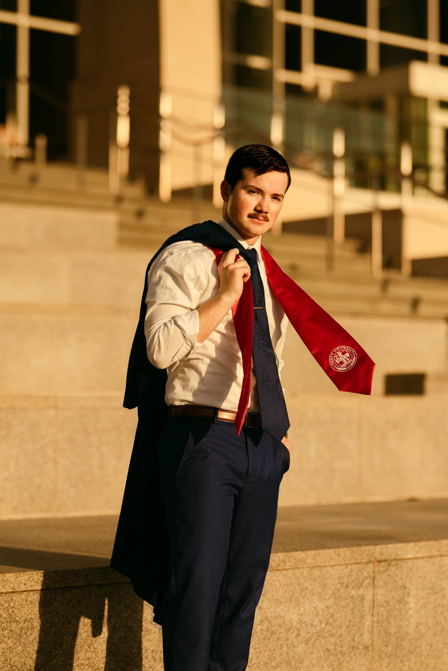 Graduate in formal attire poses near brick columns during golden hour.