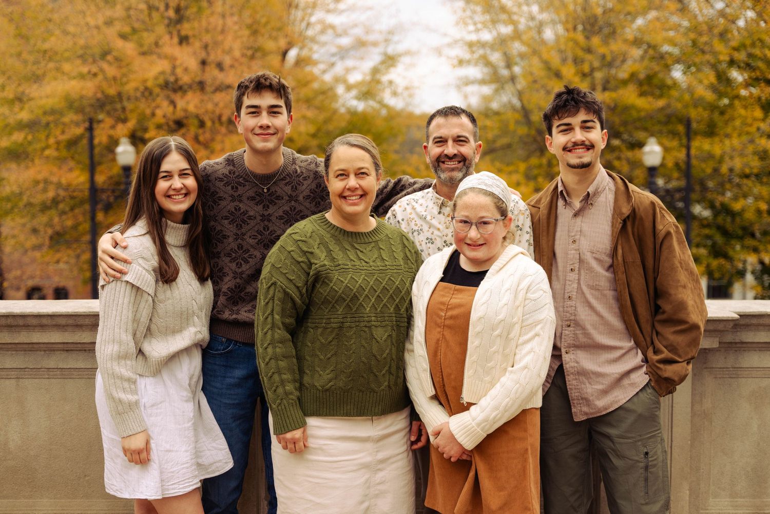 Family group wearing coordinated autumn sweaters poses together outdoors with fall foliage in background.