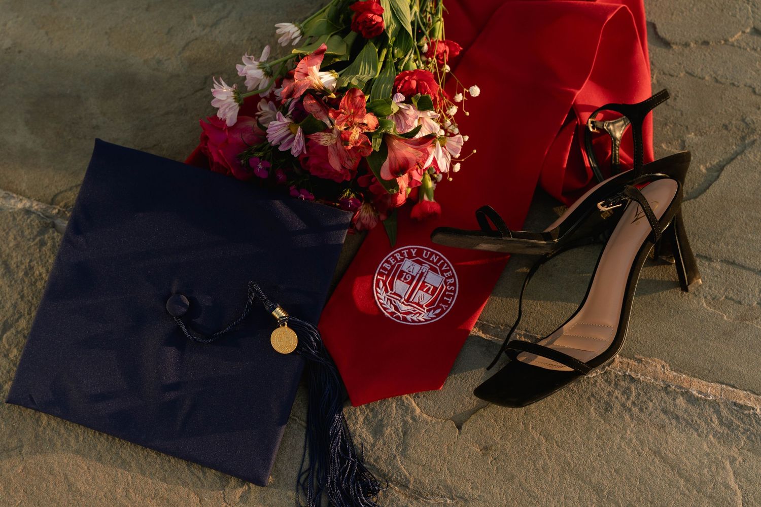 Graduation cap, red hood, diploma, heels, and flowers arranged on stone steps for a university commencement celebration.