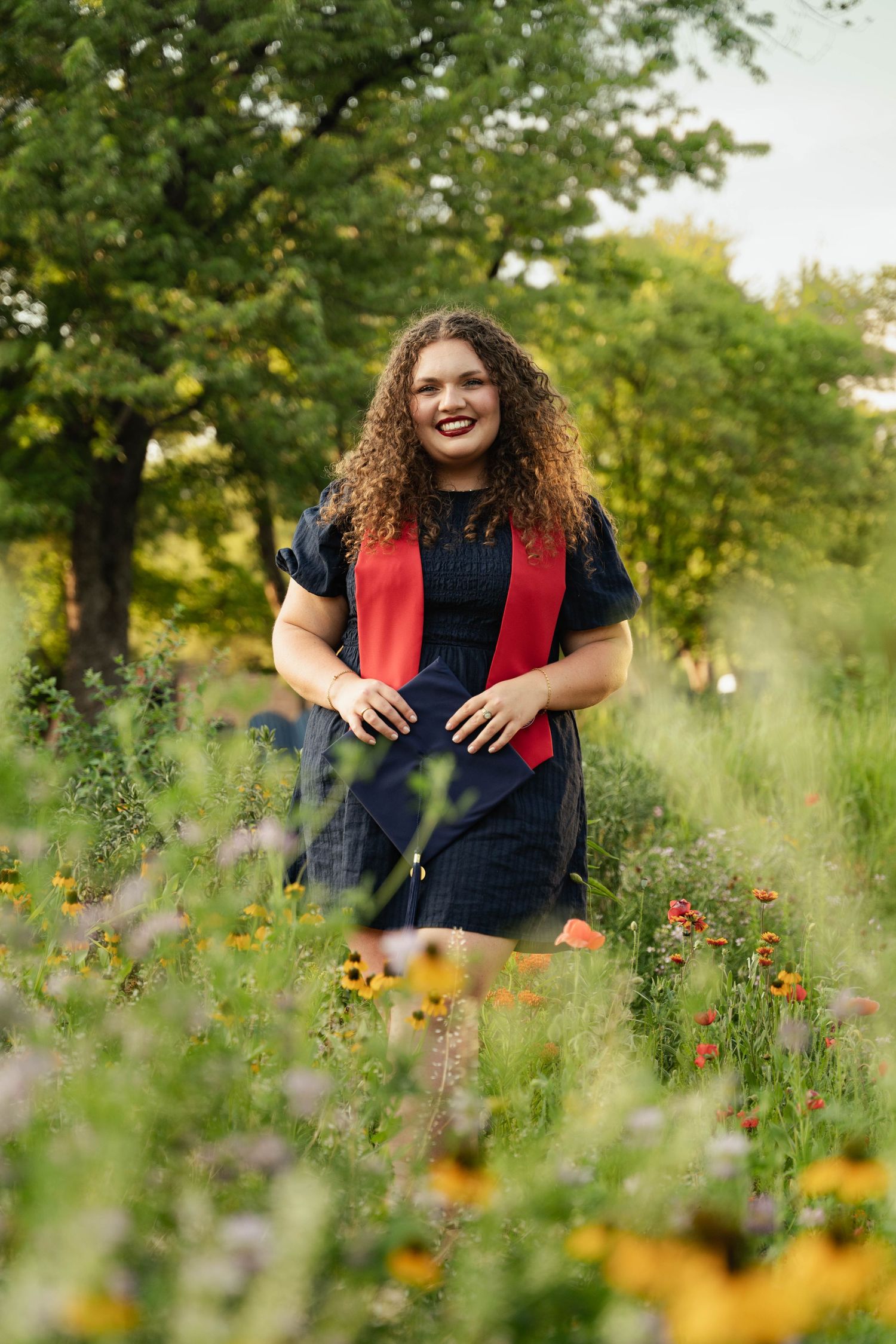 Smiling graduate in black dress with red sash stands among wildflowers in a lush green garden.