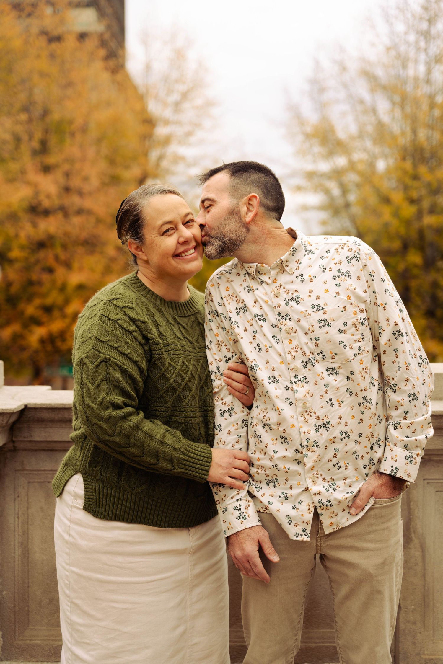 A couple wearing fall sweaters embraces and laughs together against autumn foliage.