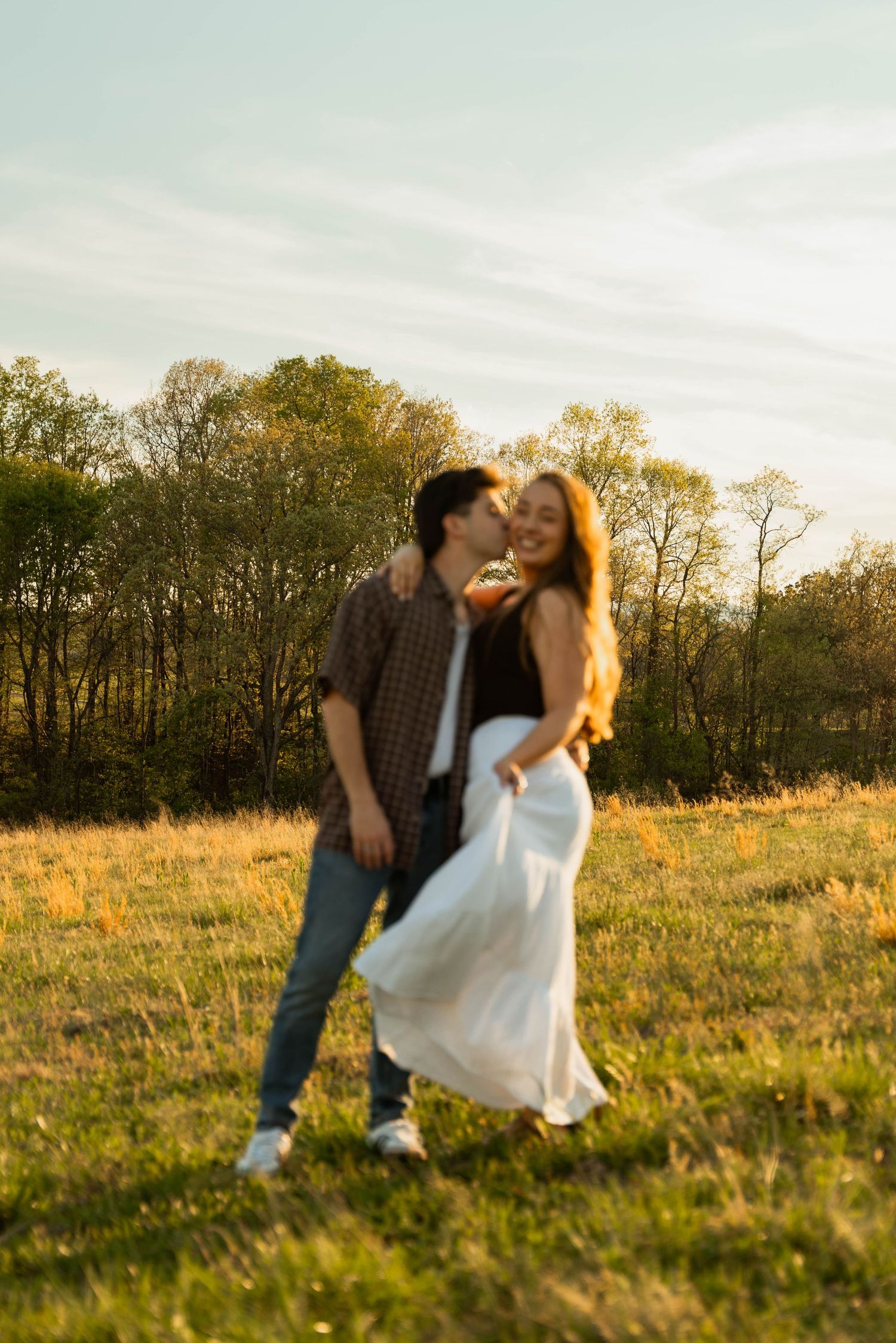 A couple shares a romantic kiss in a sunlit meadow, with the woman wearing a flowing white skirt and trees in the background.