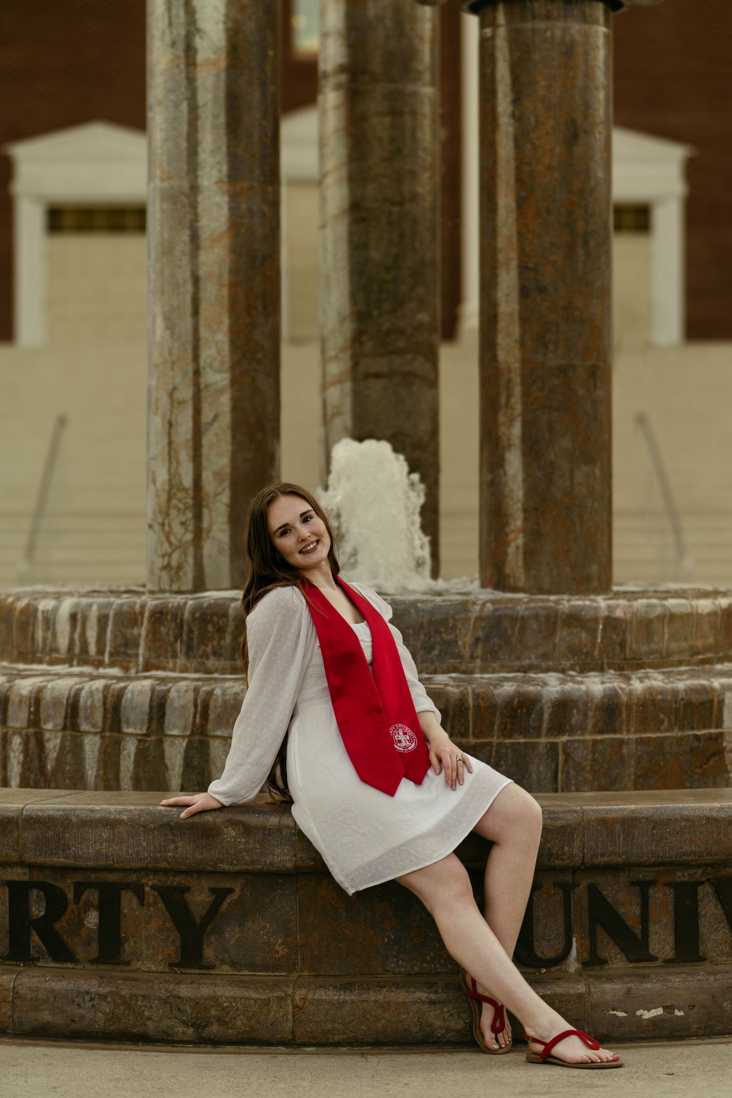 A person in a white dress and red scarf poses elegantly against stone columns in an architectural setting.