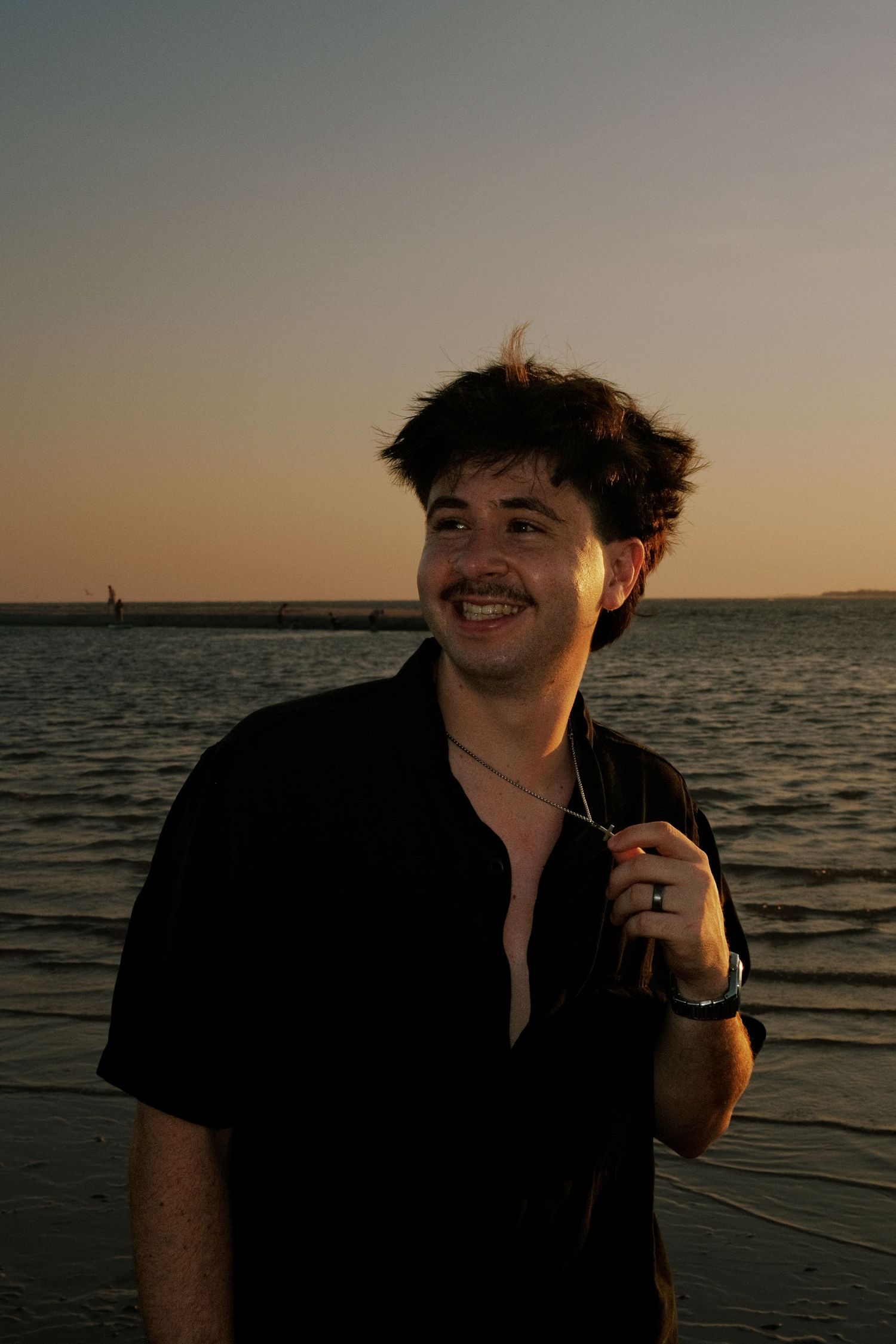 Young man with mustache smiling at sunset beach, wearing black shirt and necklace, golden hour lighting.