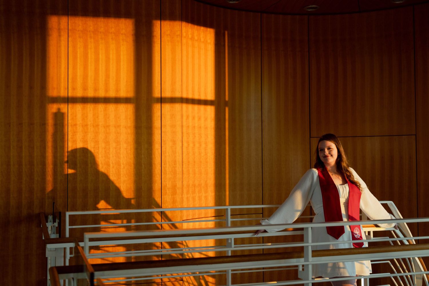 A woman in a red dress stands on a balcony, silhouetted against the warm glow of the sunset.