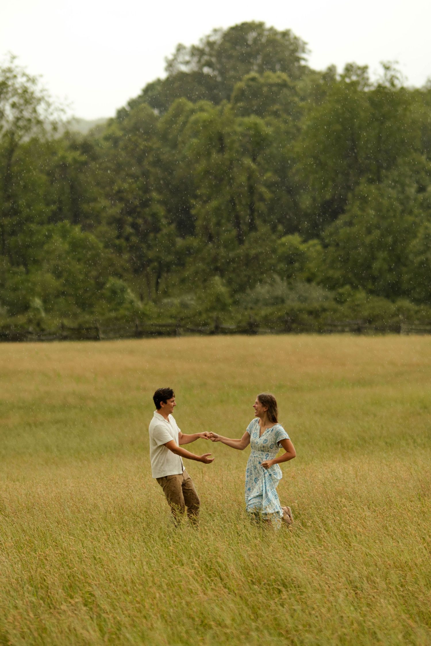 A couple holds hands and dances together in a lush green meadow field surrounded by trees.