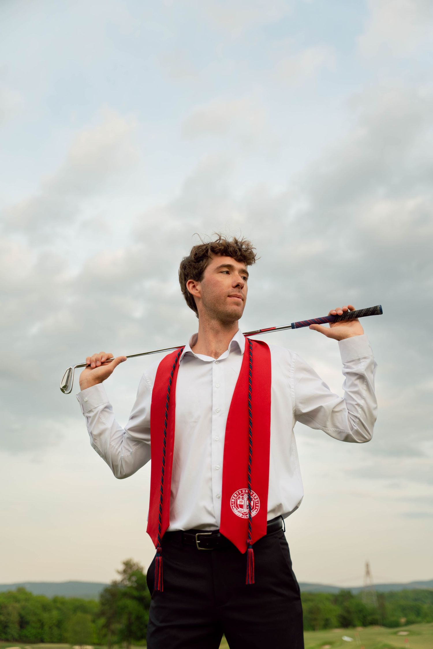 Graduate in white shirt and red stole poses outdoors against cloudy sky.