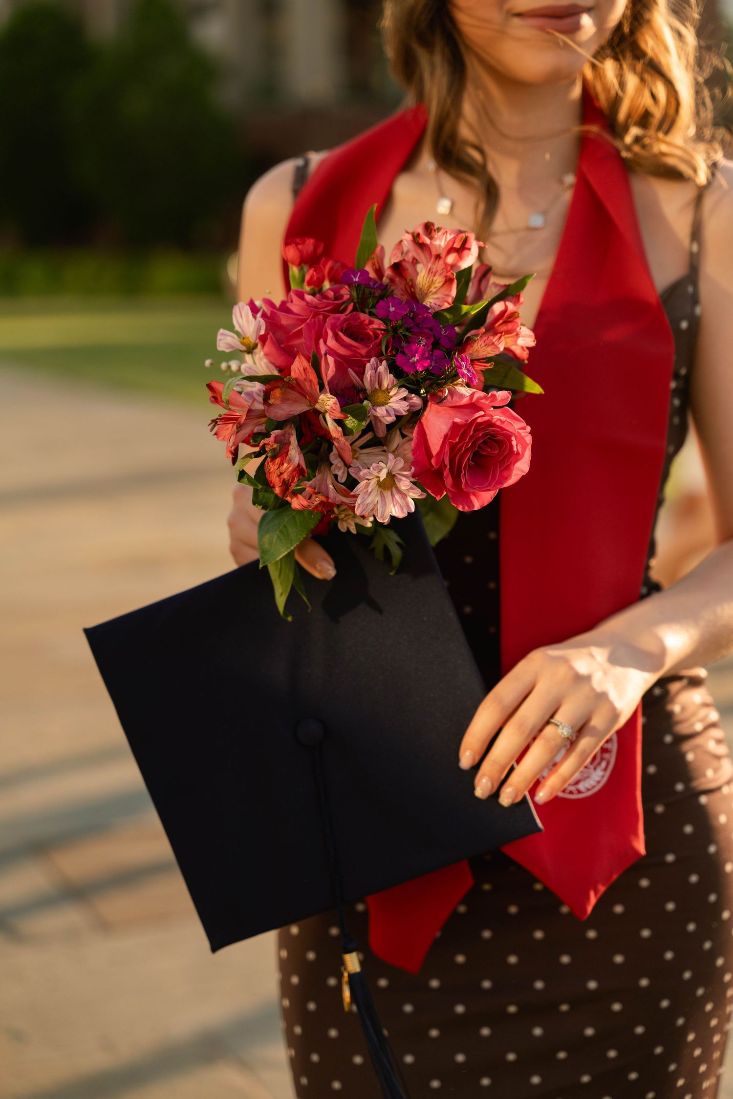 Graduate holding black mortarboard cap and vibrant pink floral bouquet, wearing red sash and polka dot dress outdoors.