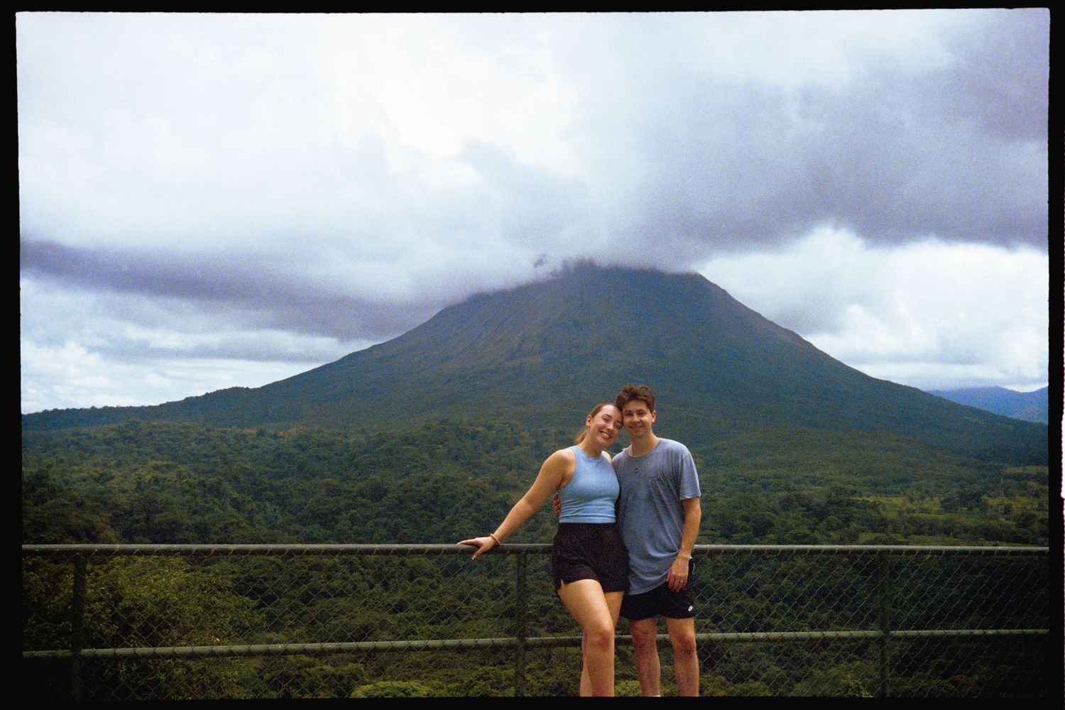 A couple standing in front of a lush, green forest and a large, volcanic mountain in the background.