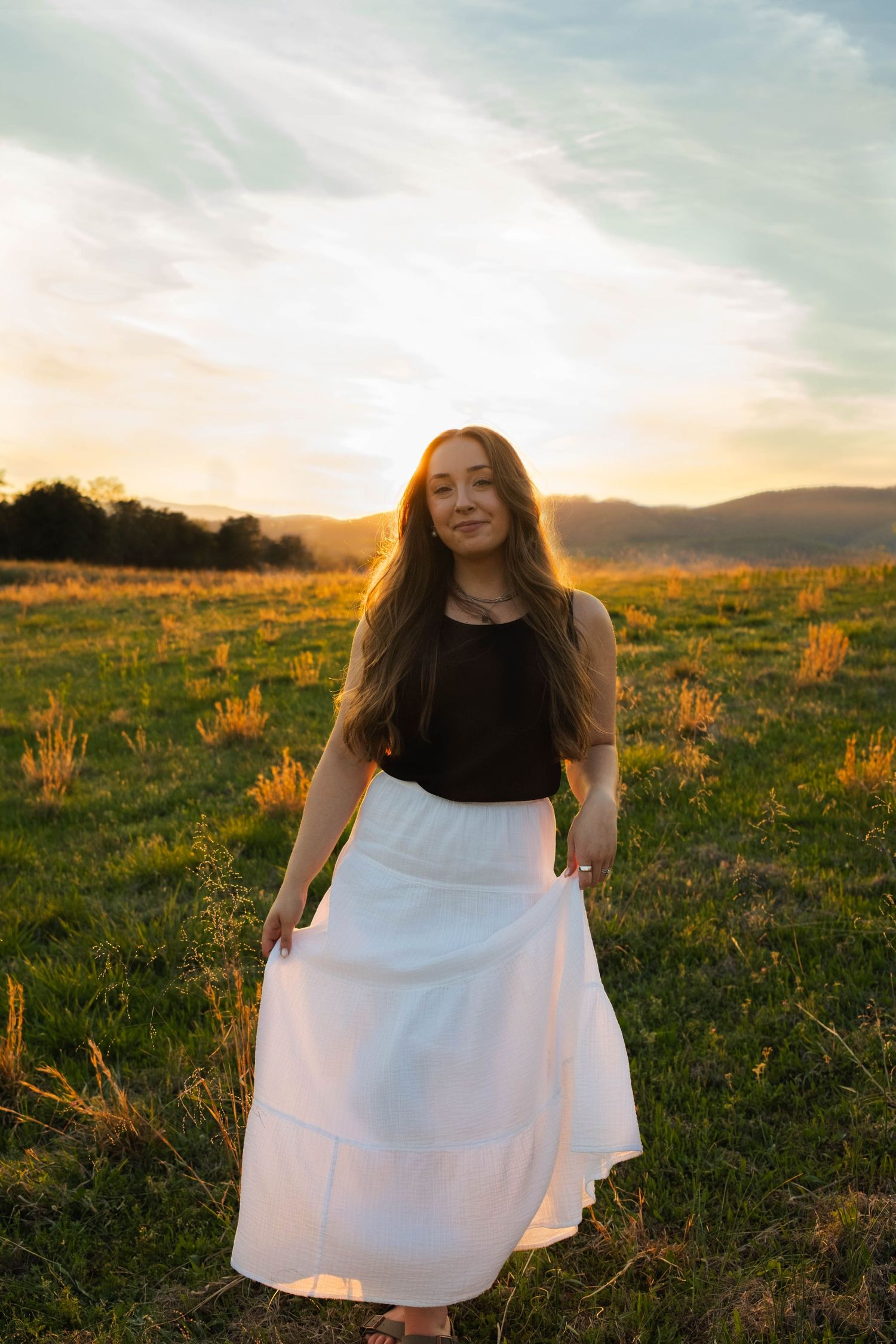 Young woman in black top and white maxi skirt standing in golden field at sunset with mountains in background.