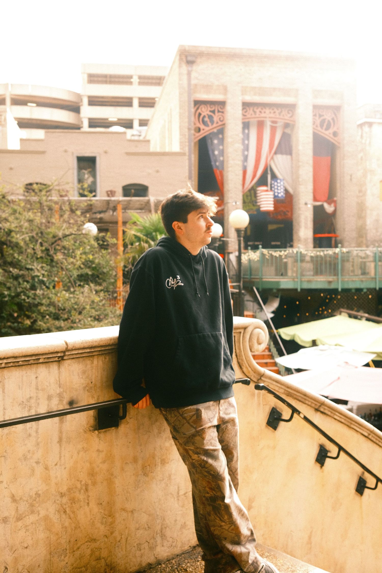 A young man wearing a black jacket stands on a wooden platform, with buildings and flags visible in the background.