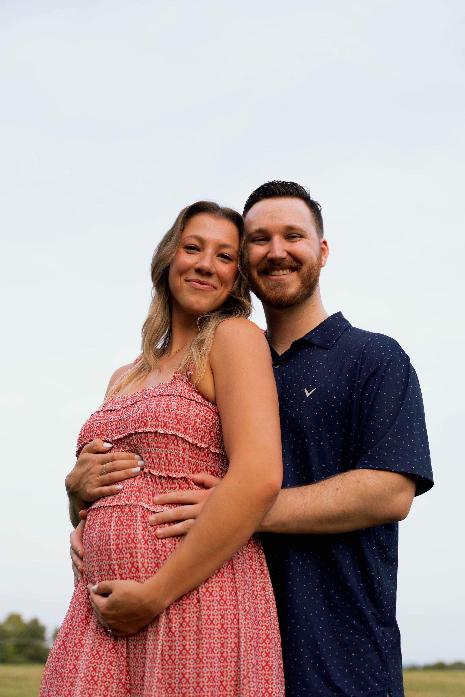 A pregnant woman wearing a pink dress is embraced by a smiling man in a navy blue shirt in an outdoor field setting.